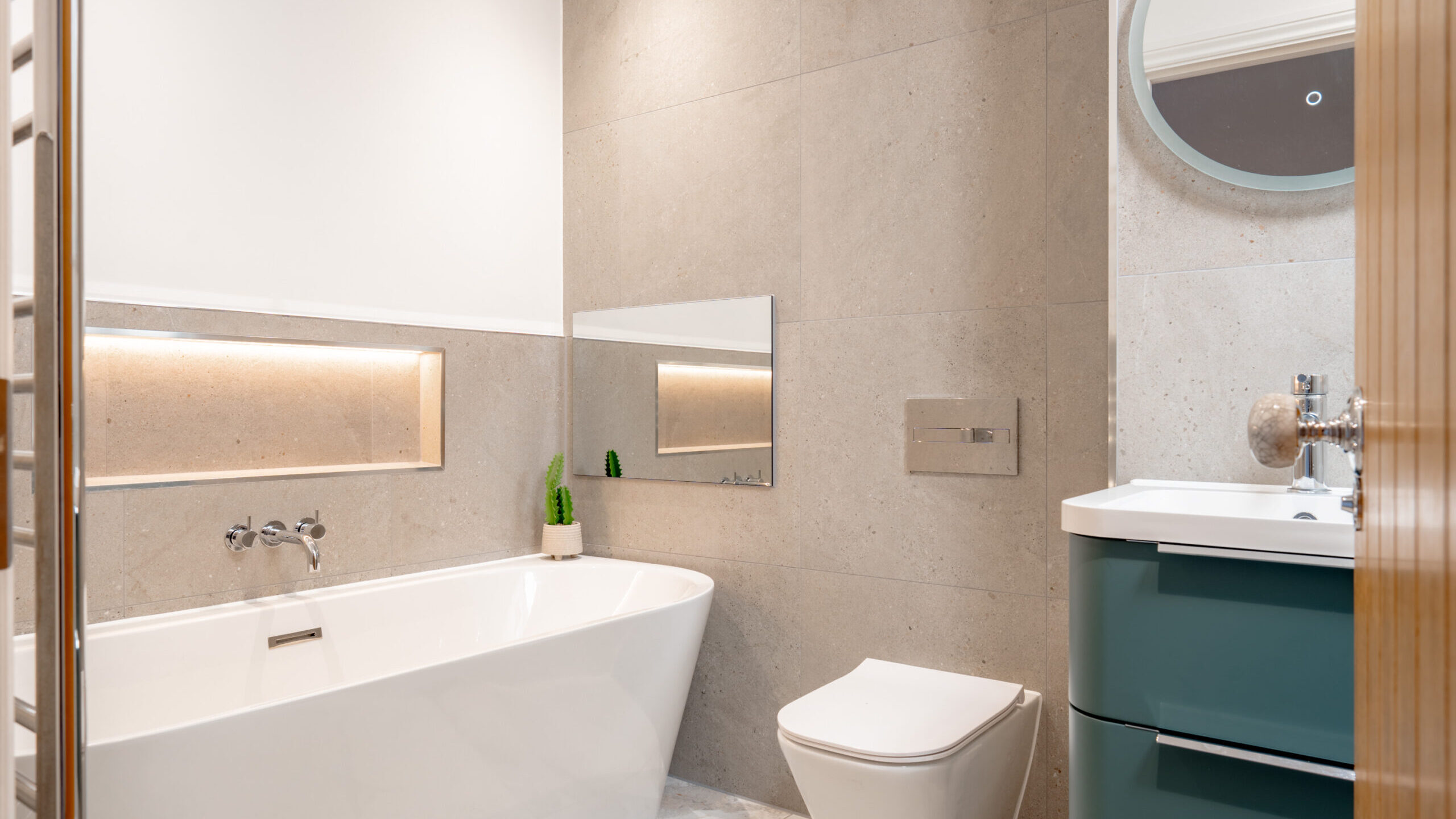 Modern bathroom with a white bathtub, wall-mounted toilet, floating sink cabinet with green drawers, round mirror, and minimalist decor including a small potted plant and built-in shelf lighting.