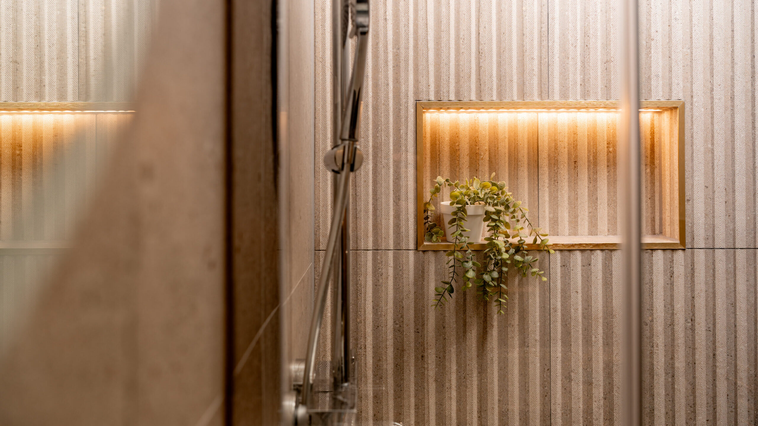 Modern shower with ribbed beige wall tiles, a built-in illuminated shelf holding a small potted green plant, and a metallic showerhead visible in the foreground.