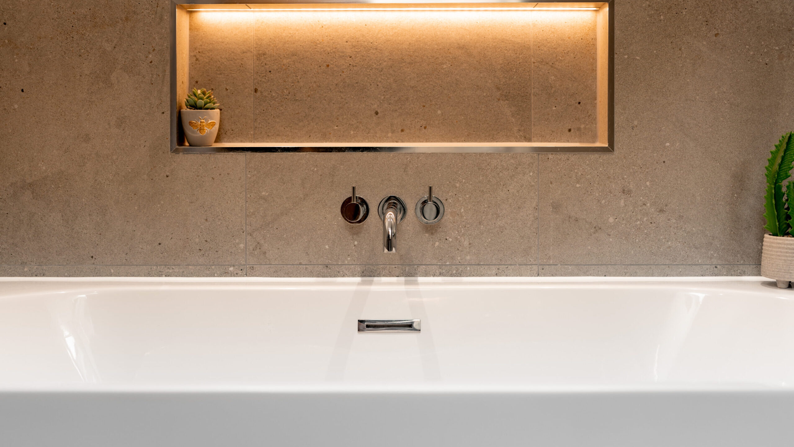 A modern bathtub with a gray stone backsplash, built-in shelf with warm lighting, a small potted plant on the left, and a green plant on the right side of the tub. Two round faucet handles and a spout are centered above the tub.