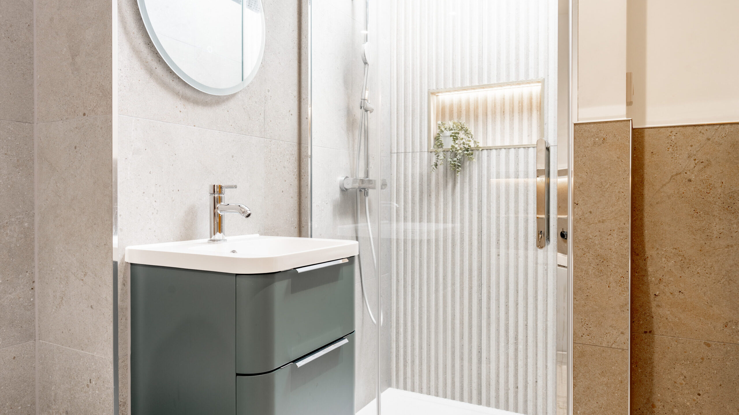 Modern bathroom with a round mirror above a sleek vanity, adjacent to a glass-enclosed shower with vertical tile, built-in shelf, and a small green plant. Clean, neutral tones and minimalist design.