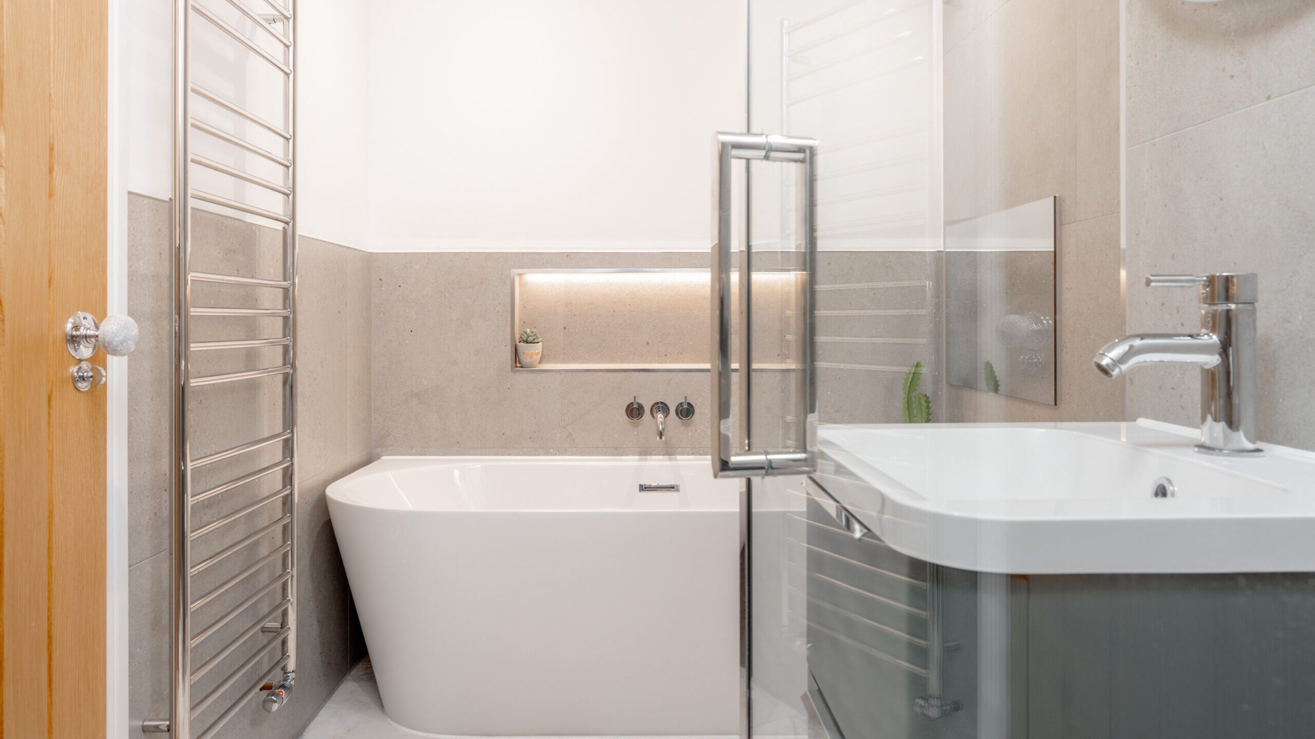 Modern bathroom with a white freestanding tub, a glass shower door, a sleek sink with chrome faucet, a wall-mounted towel radiator, and a small shelf with decor above the tub. The decor is minimal and the space is bright.