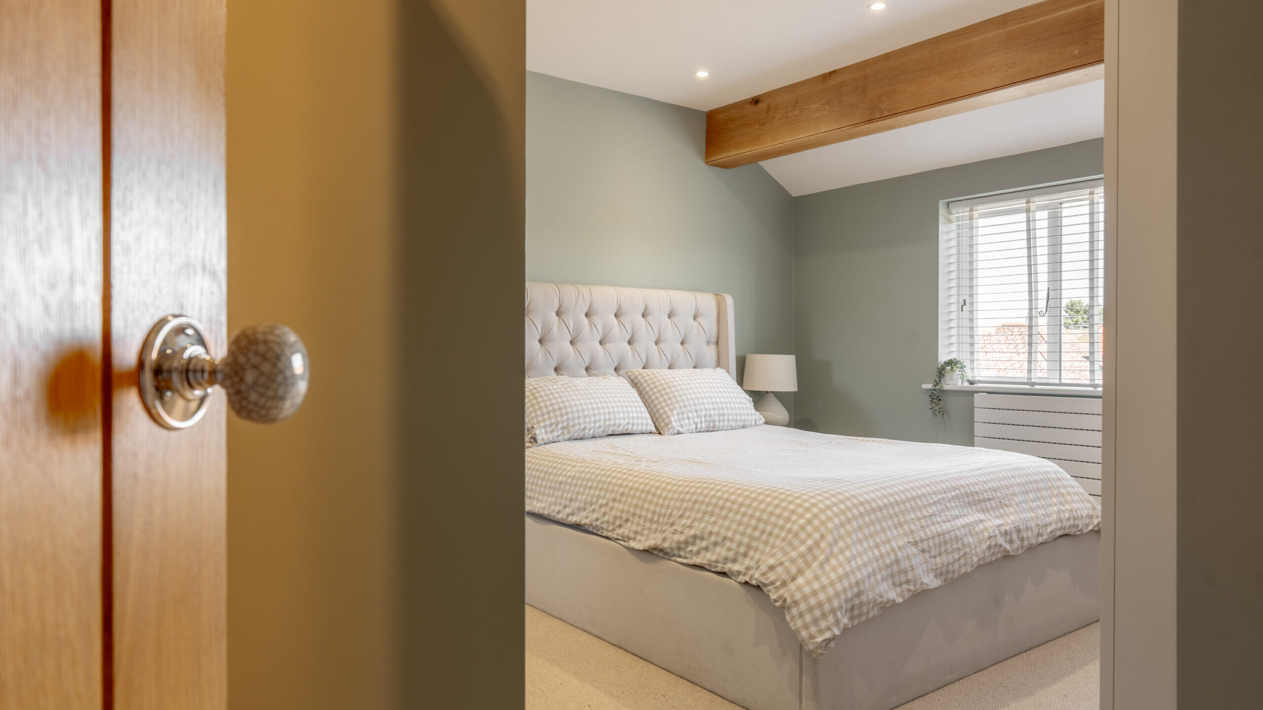 A cozy bedroom with a tufted headboard, neatly made bed with checkered bedding, a bedside lamp, a window with blinds, a potted plant, and a wooden ceiling beam, viewed from a doorway.