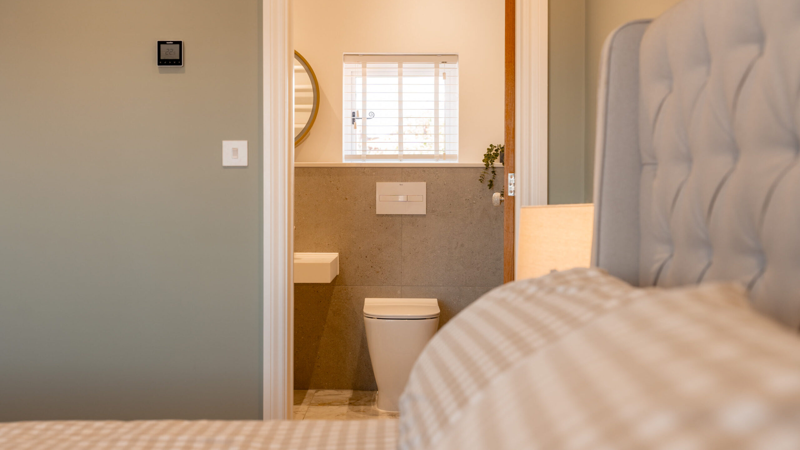 A view from a bedroom with a checkered bedspread, looking into a small bathroom with a toilet, round mirror, and window with white blinds. The bathroom walls are light-colored and a lamp is visible beside the bed.