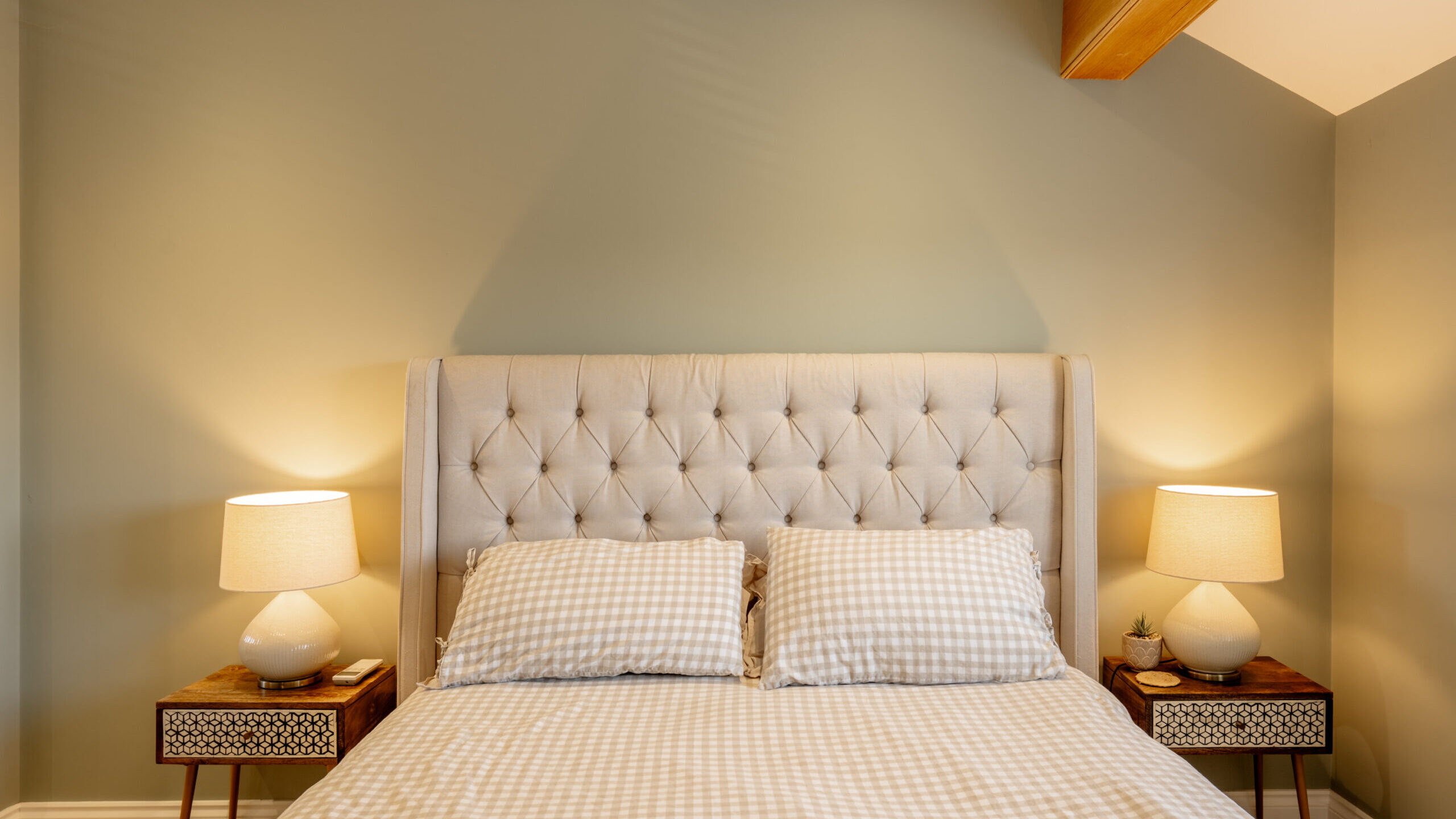 A neatly made bed with a tufted beige headboard, two checked pillows, and matching bedding. Two nightstands with lamps and small decor items are on each side, against a light green wall.