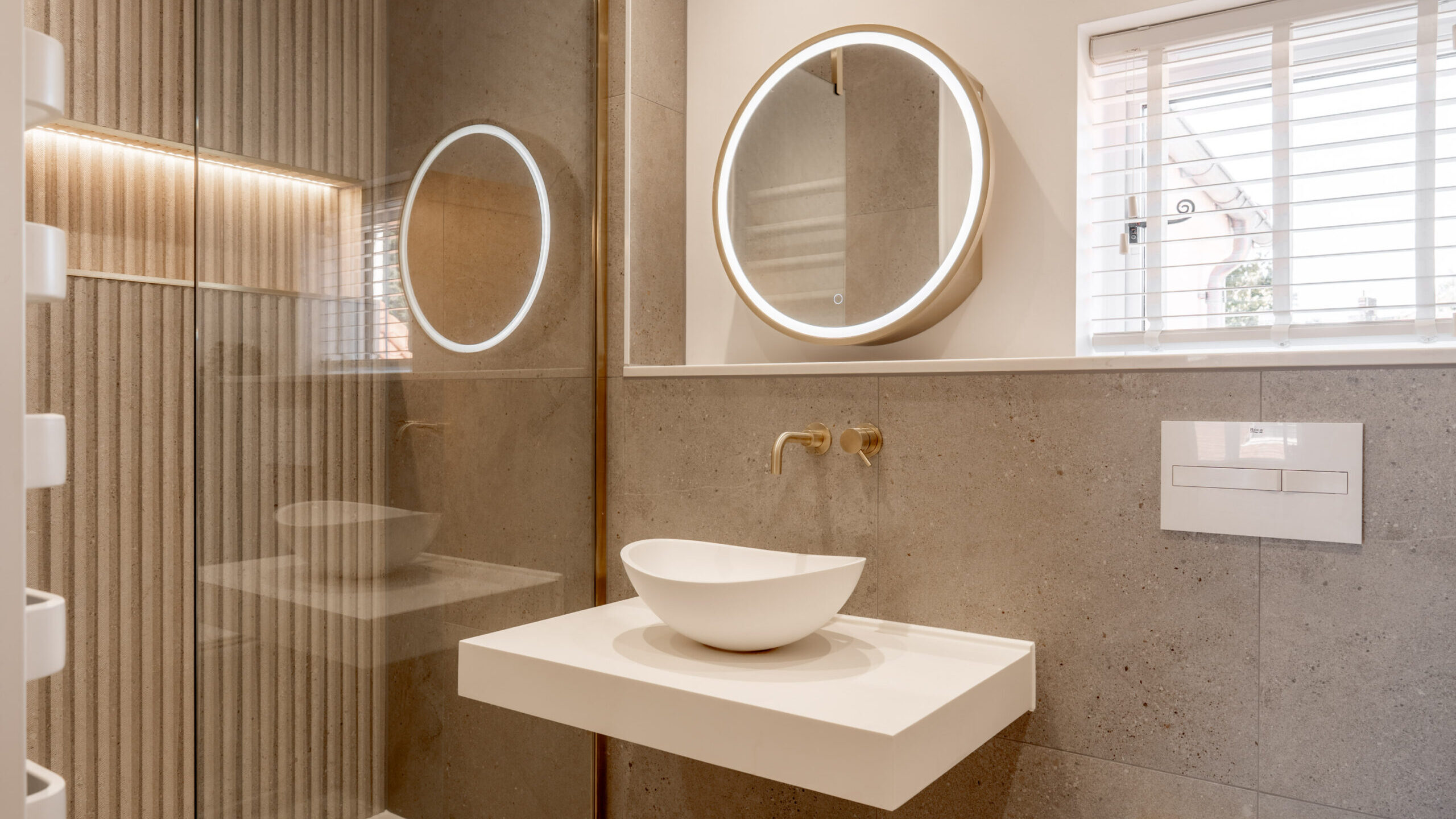 Modern bathroom with a round backlit mirror, vessel sink on a floating white vanity, wall-mounted gold faucet, glass shower enclosure, and beige tiled walls. A small window with white blinds is on the right.