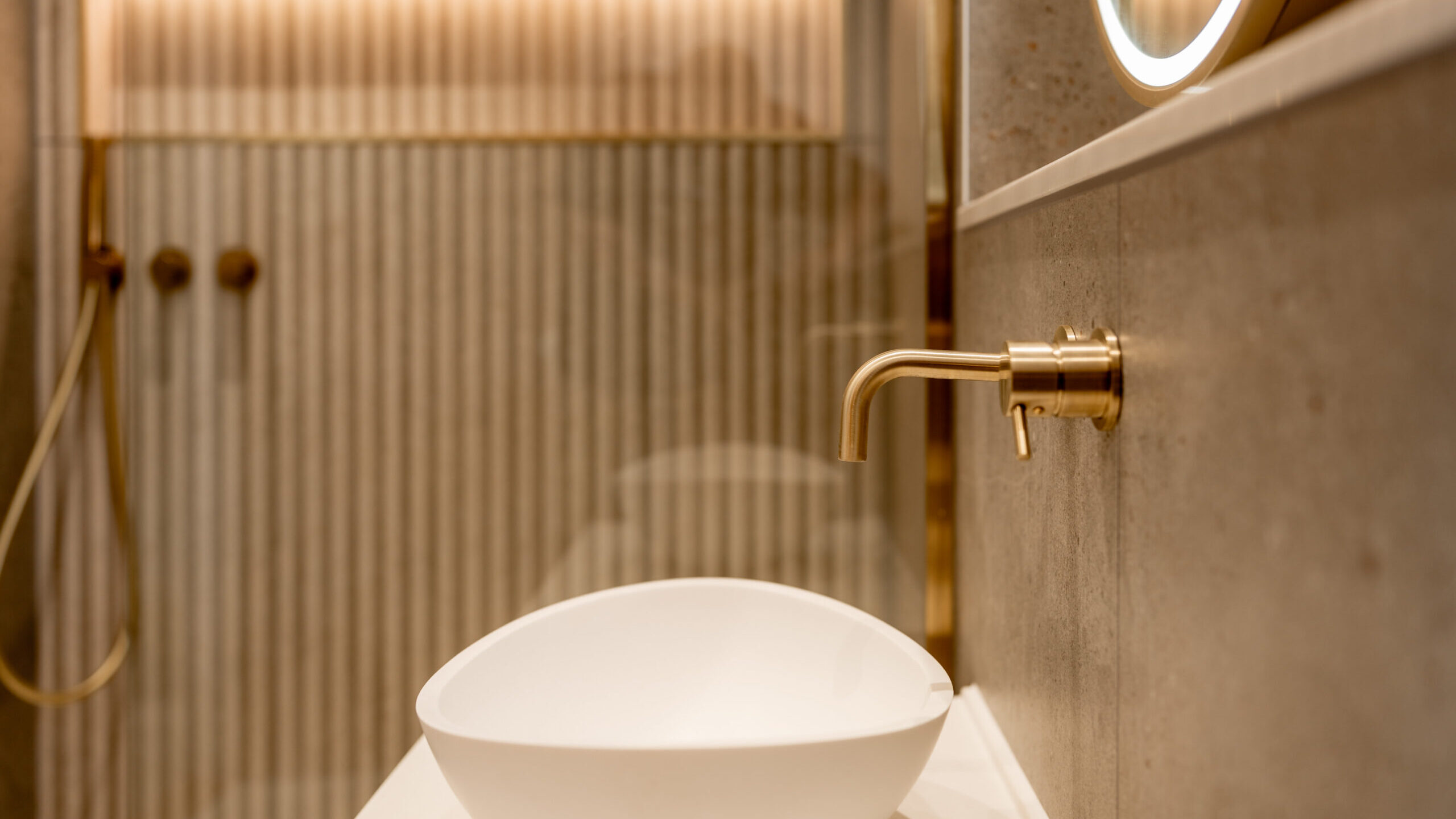 A modern bathroom with a white vessel sink, a gold wall-mounted faucet, and a round illuminated mirror. The background features a textured, striped wall with gold shower fixtures and soft, warm lighting.