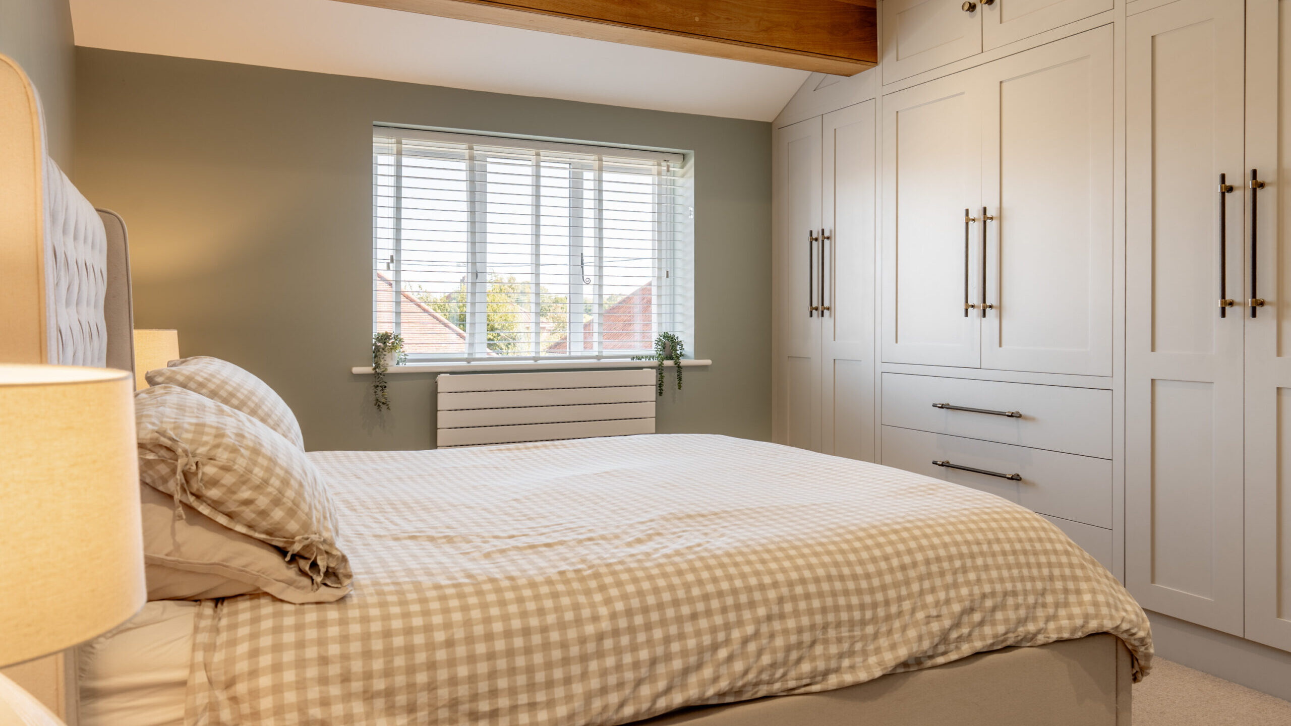 A cozy bedroom with a neatly made bed in plaid bedding, white built-in wardrobes with black handles, a large window with blinds, and a wooden ceiling beam above.