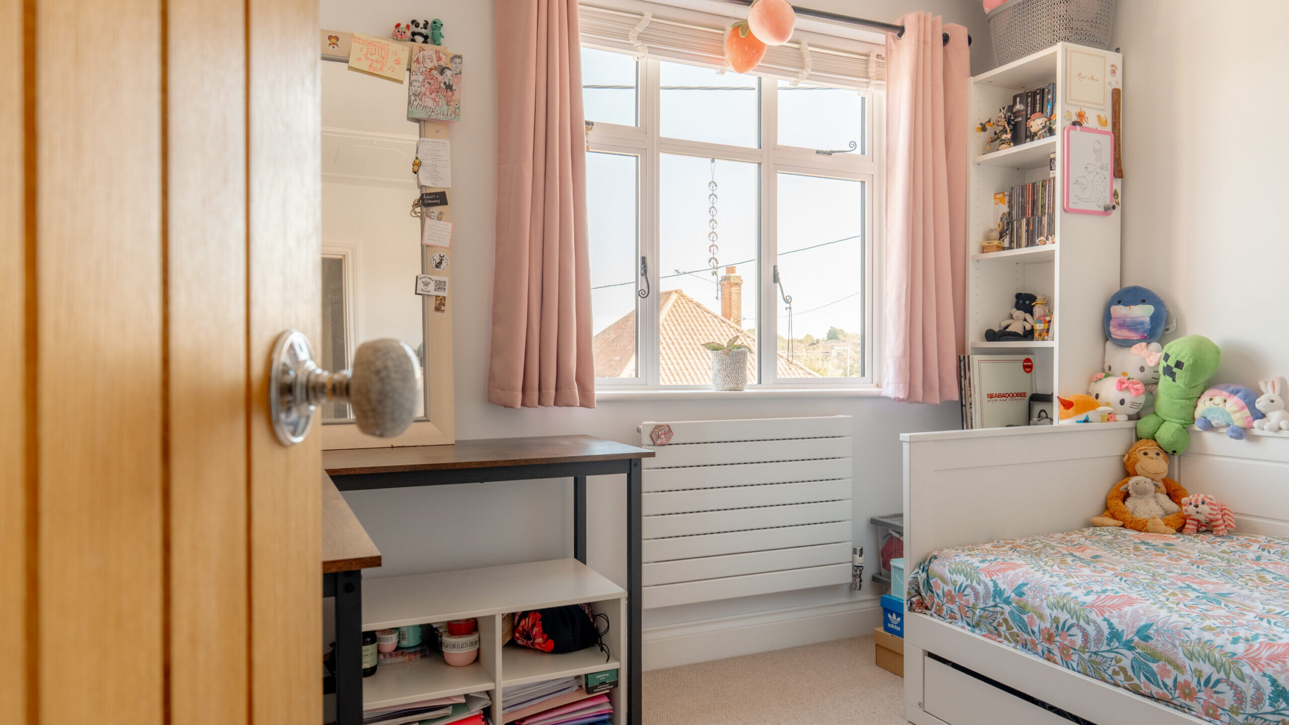 A cozy, tidy bedroom with light pink curtains, a window, a bed with colorful stuffed animals, a floral bedspread, a desk with shelves, and a mirror on the wall. Sunlight fills the room.