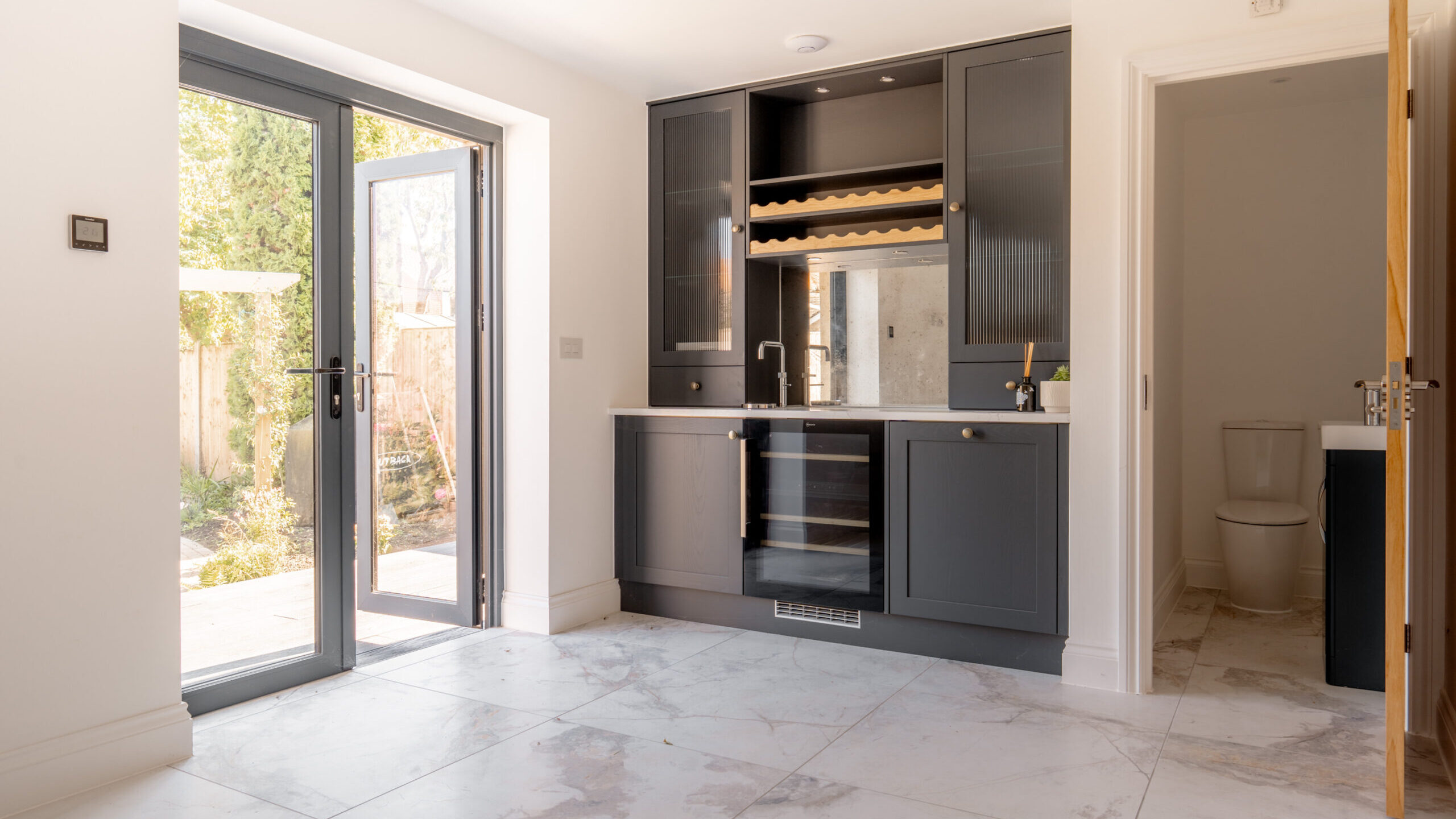 Modern kitchen with gray cabinets, a wine rack, glass doors to a garden, marble floor tiles, and an open door leading to a bathroom with a visible toilet. Bright natural light fills the space.