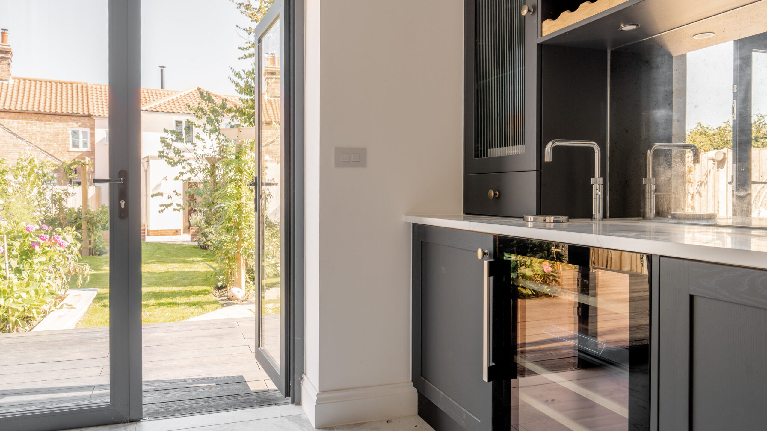 Modern kitchen with dark cabinets, a sink, and glass-fronted wine cooler. Glass doors open to a sunlit garden with green grass, flowers, and an outdoor seating area.