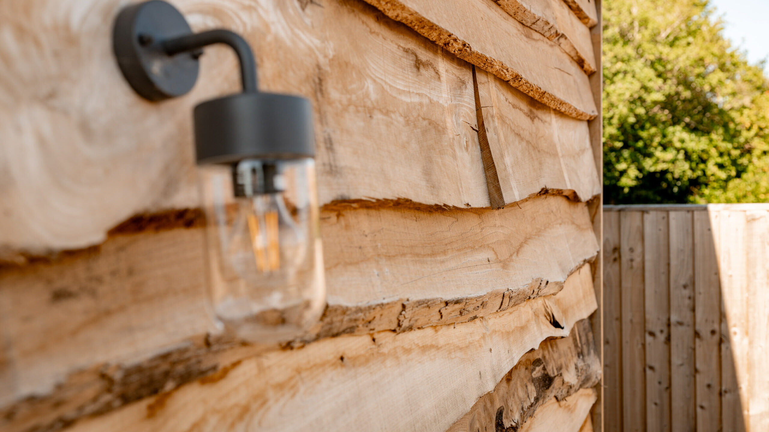 A black outdoor wall light fixture is mounted on a rustic wooden exterior wall with natural, uneven planks. A wooden fence and greenery are visible in the background.