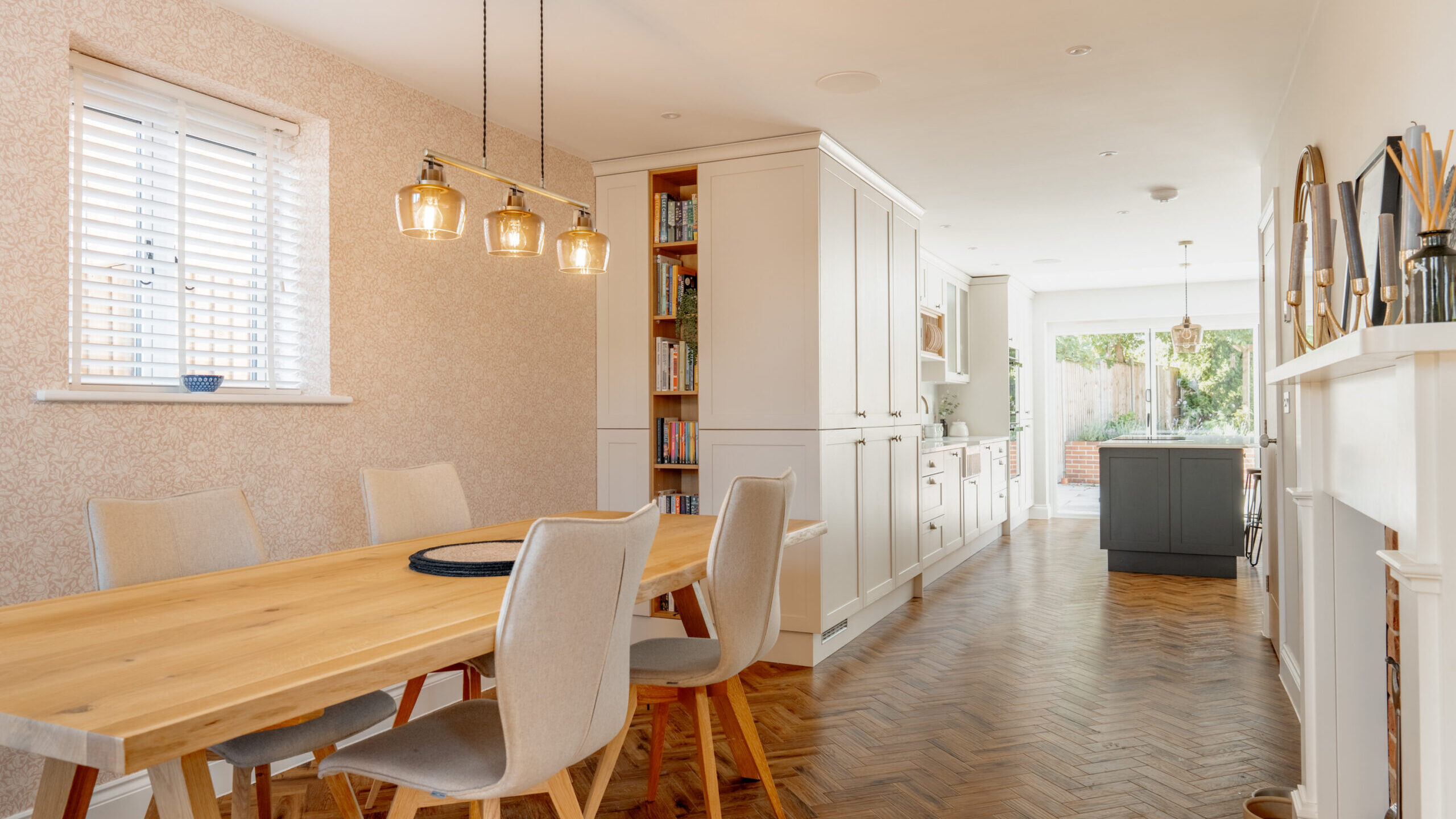 A modern dining area with a wooden table and six beige chairs, pendant lights above, and a view into a bright kitchen with white cabinets and parquet flooring. Sunlight streams in through windows at the back.