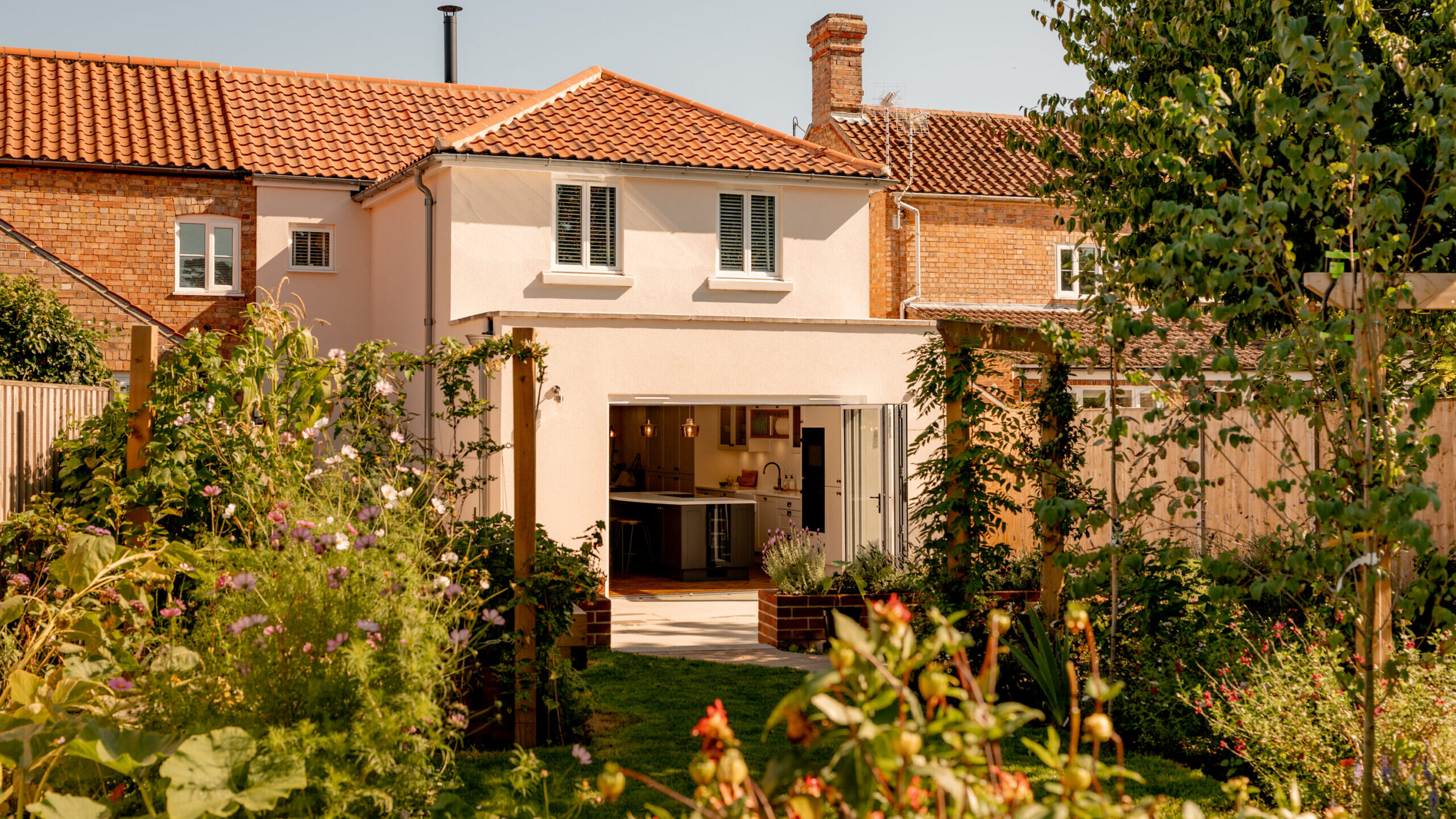 A modern two-story house with cream walls and reddish-brown tiled roof, large open patio doors leading to a bright kitchen, surrounded by a lush, colorful garden on a sunny day.