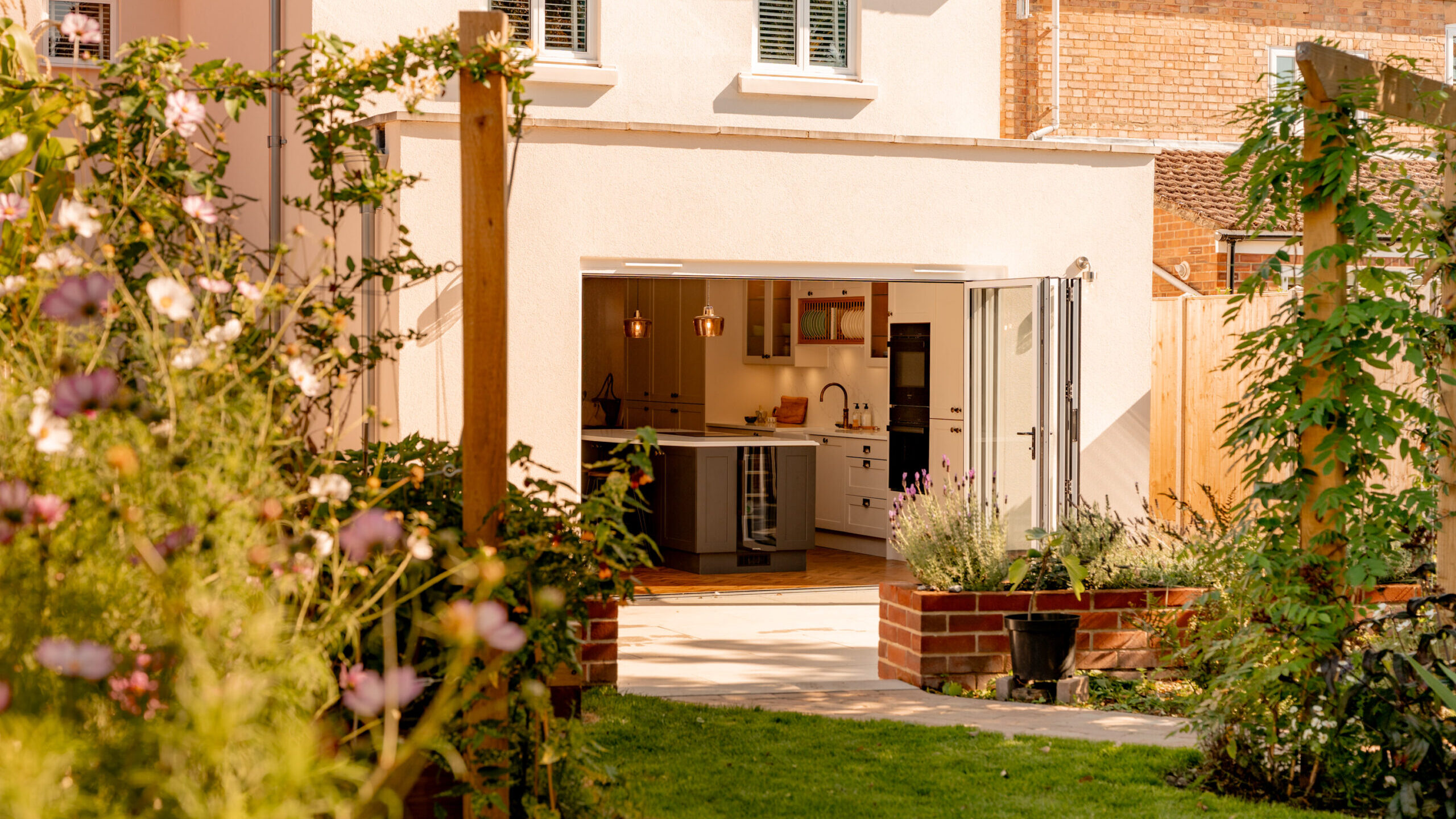 View of a modern home’s open kitchen and patio, seen from a lush garden with green grass, flowers, and climbing plants on wooden posts, on a sunny day.
