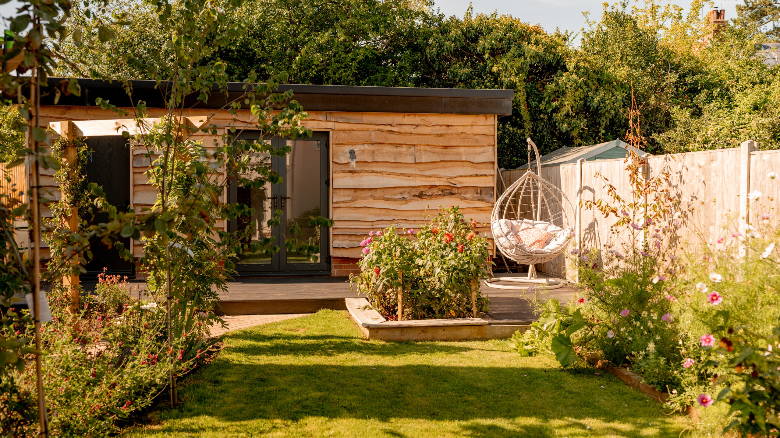 A wooden garden shed with glass doors sits in a lush backyard with green grass, flowering plants, and an egg-shaped hanging chair near a wooden fence under a sunny sky.