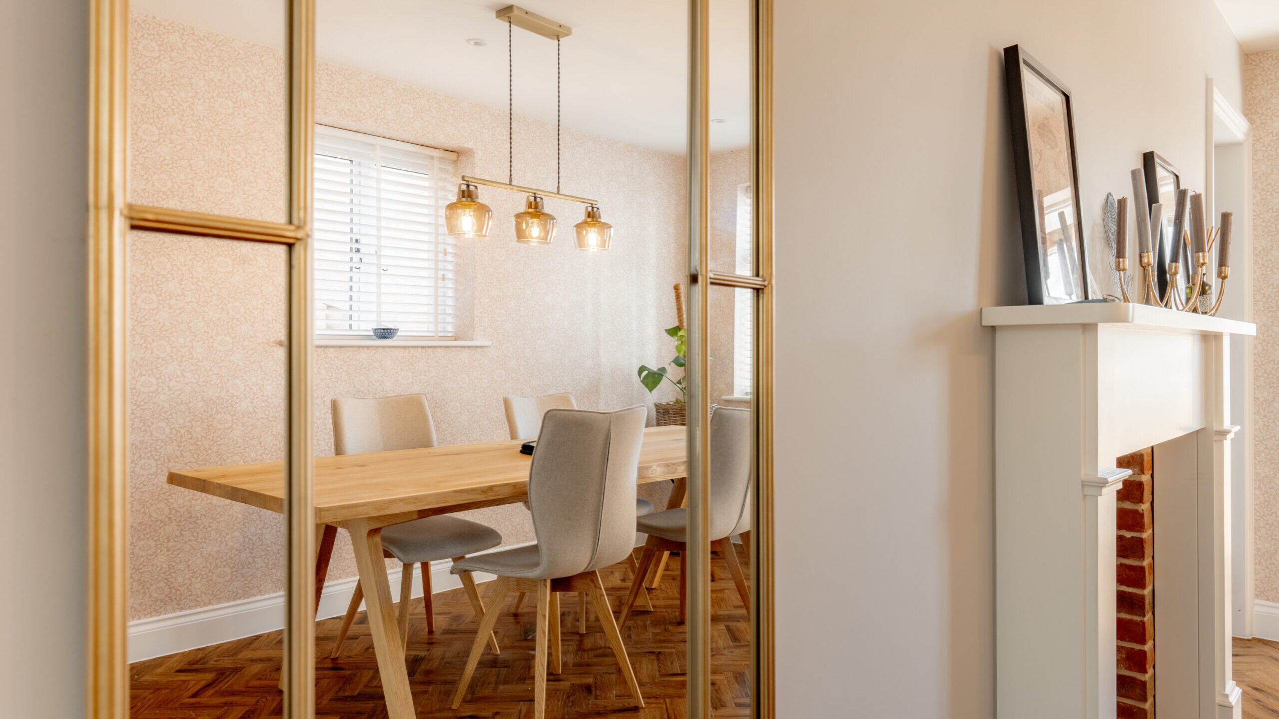 A modern dining room with a wooden table, beige chairs, and a three-light pendant fixture, reflected in a large wall mirror. The room has parquet flooring and a white fireplace with decor.