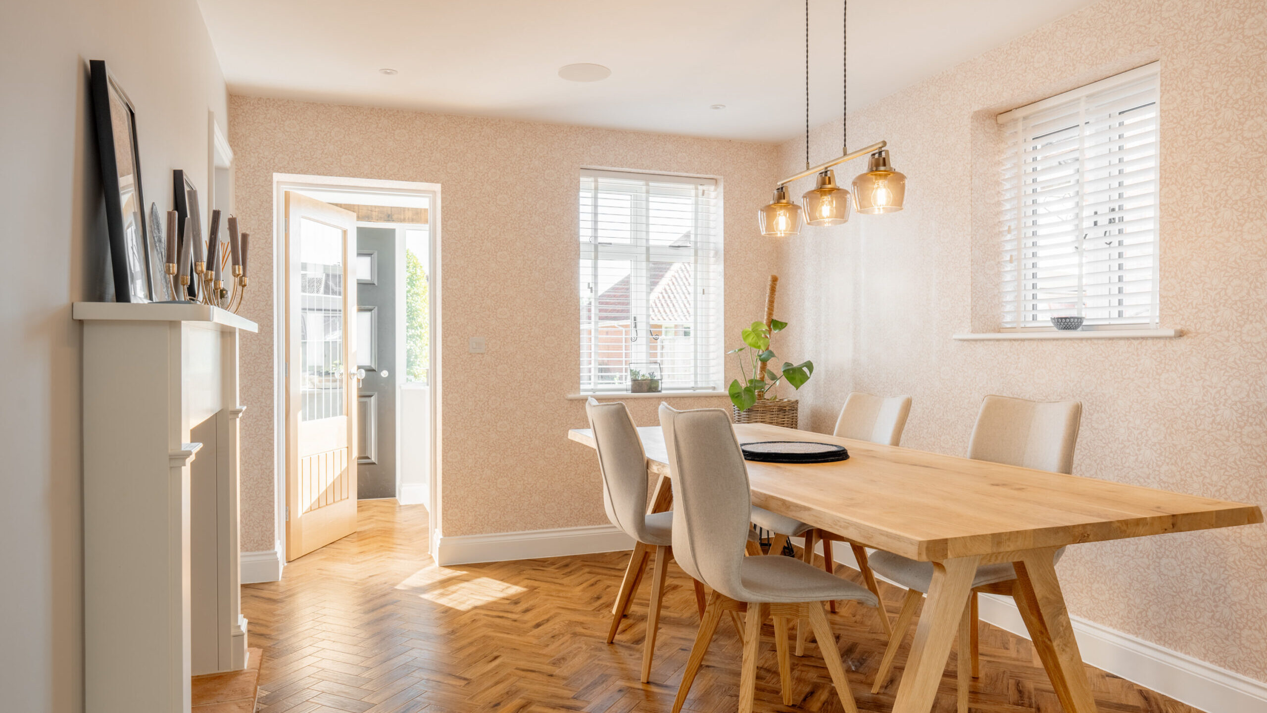 Bright dining room with a wooden table, six upholstered chairs, light wood herringbone floor, pendant light fixture, wall art on a mantel, and windows with white blinds letting in natural light.