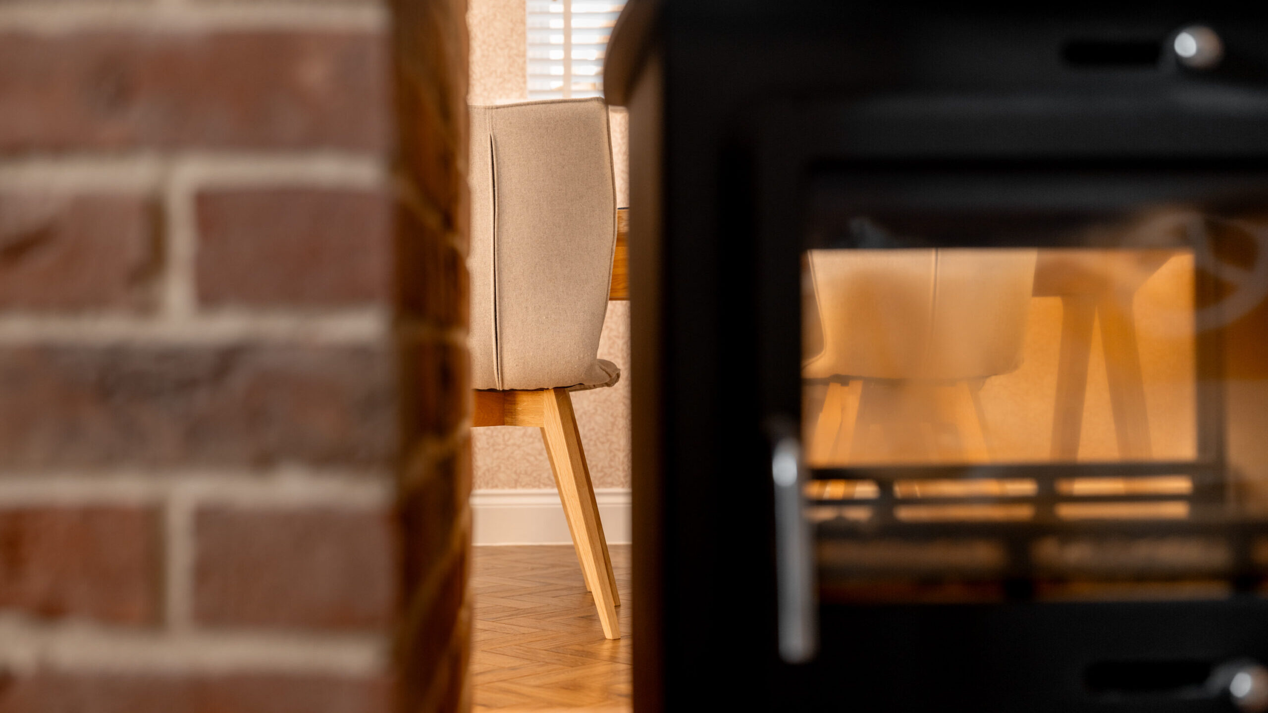 Close-up of a black wood-burning stove on the right, partially out of focus, with a brick wall and a beige dining chair at a wooden table in the background. White blinds cover a window in the background.