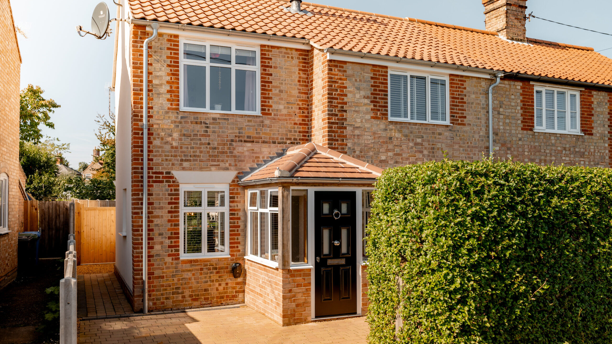 A two-story brick house with a red-tiled roof, white-framed windows, and a black front door. A trimmed green hedge lines the walkway beside the house, and a wooden gate is visible on the left.