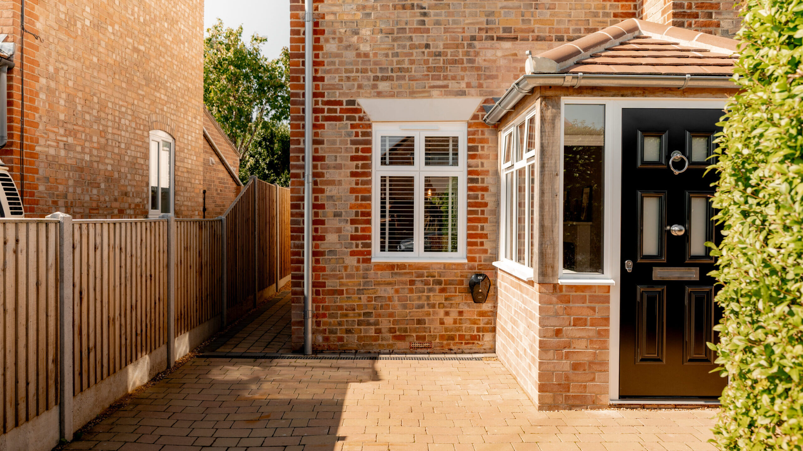 A modern brick house with white-framed windows, a black front door, and a small porch. The driveway is paved with bricks, and a wooden fence and tall green hedge border the house.