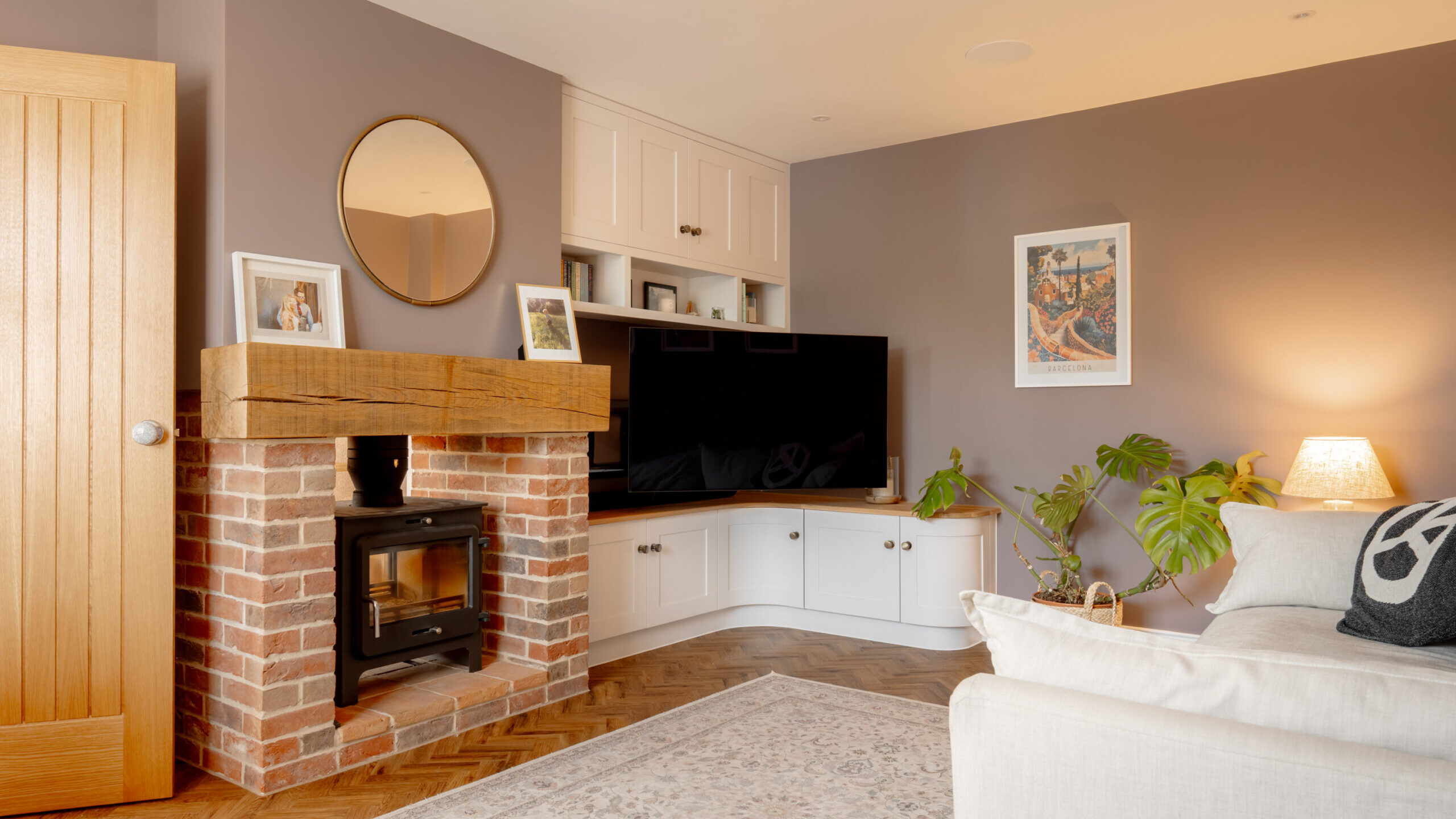A cozy living room with a wood-burning stove set in a brick fireplace, a large TV on a white media unit, a sofa, houseplants, framed pictures, a round mirror, and soft lighting from a table lamp.
