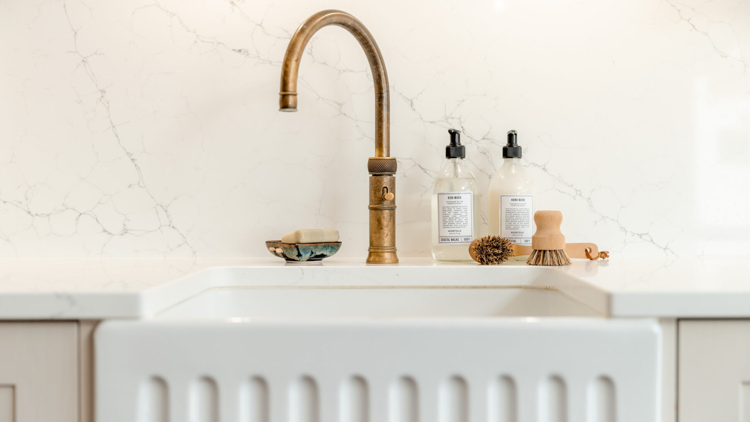 A brass kitchen faucet over a white sink with a ribbed front; on the counter are soap dispensers, a scrub brush, a wooden dish brush, and a small dish, all against a white marble backsplash.