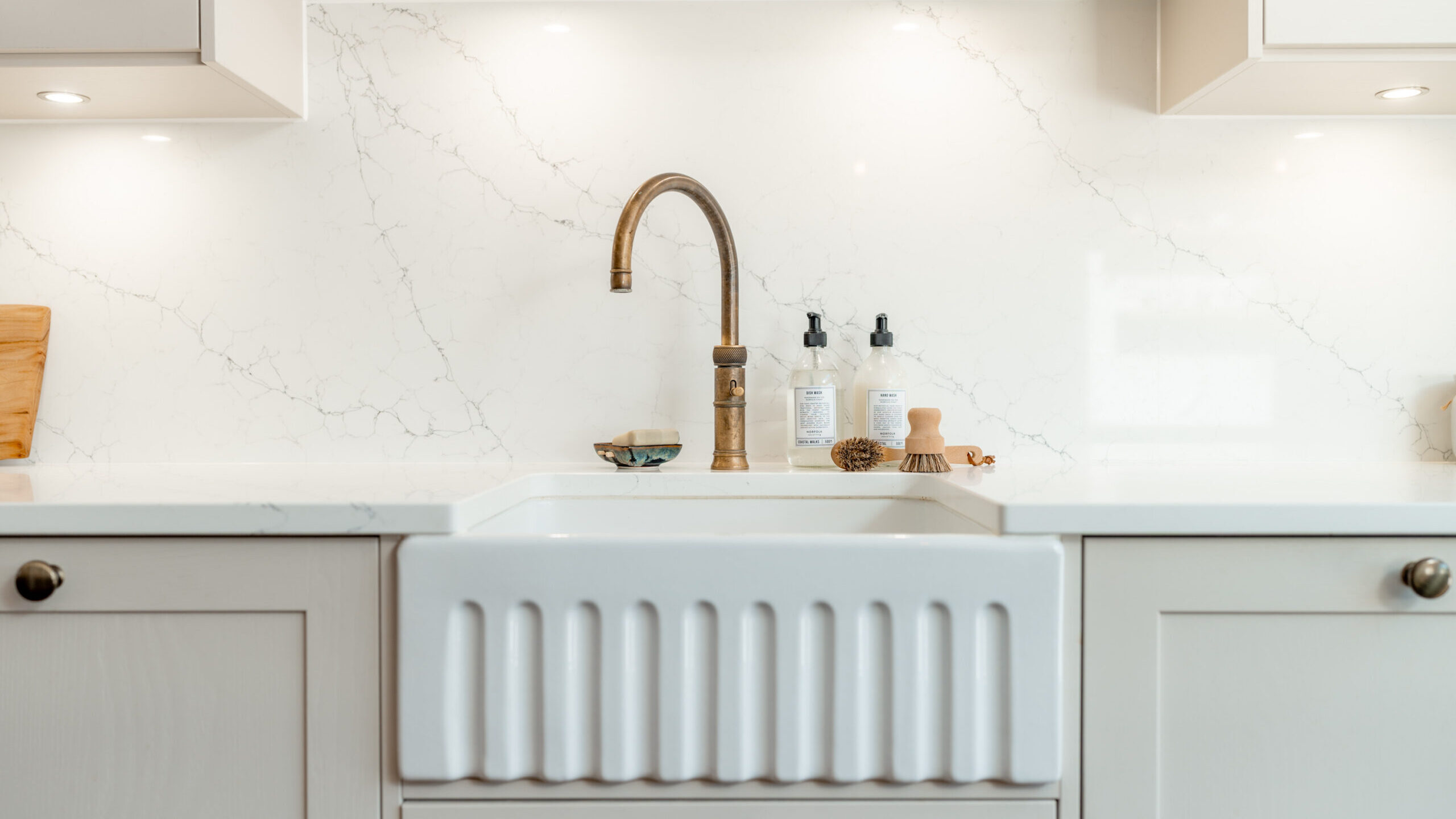 Close-up of a modern kitchen sink with a brass faucet, white countertop, and marble-patterned backsplash. Soap dispensers and a small brush are placed on the counter next to the sink.
