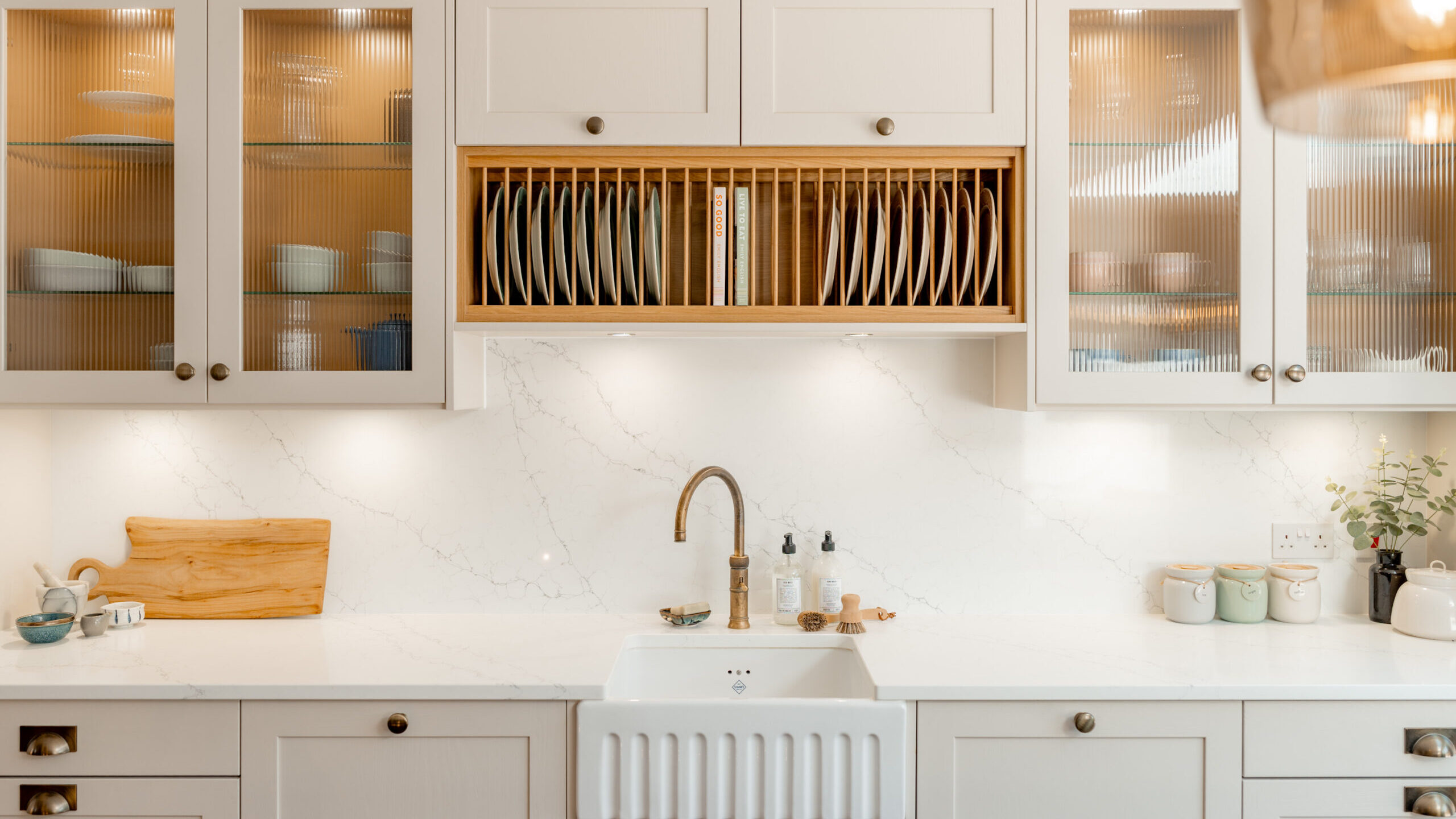 A bright, modern kitchen with white cabinets, a farmhouse sink, a brass faucet, a wooden dish rack, and glass-front cupboards displaying dishes. The countertop holds jars, soap dispensers, and a wooden cutting board.