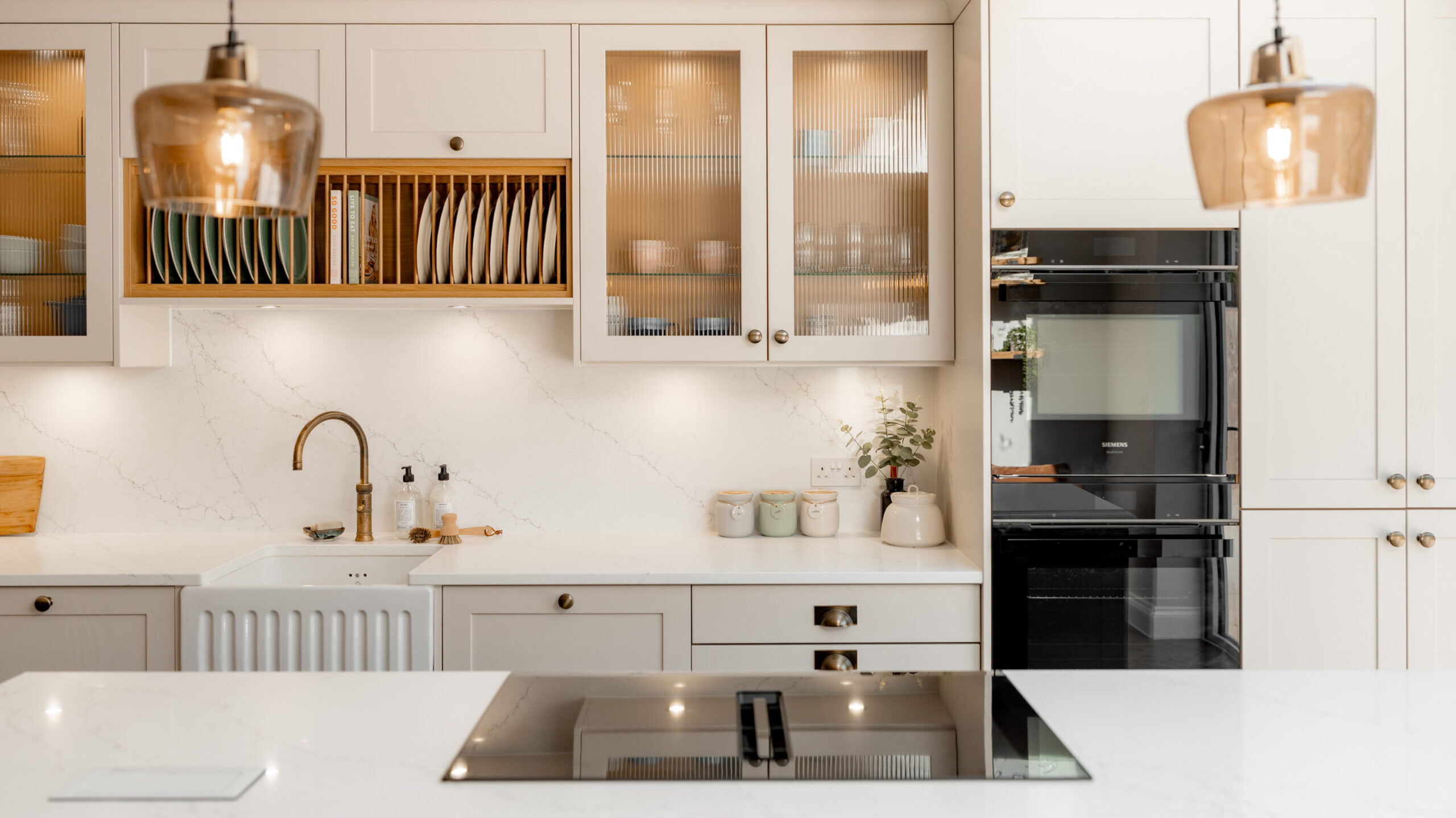 Bright, modern kitchen with white cabinets, marble countertops, a farmhouse sink, built-in oven, electric stove on an island, glass pendant lights, and neatly arranged dishes and jars.