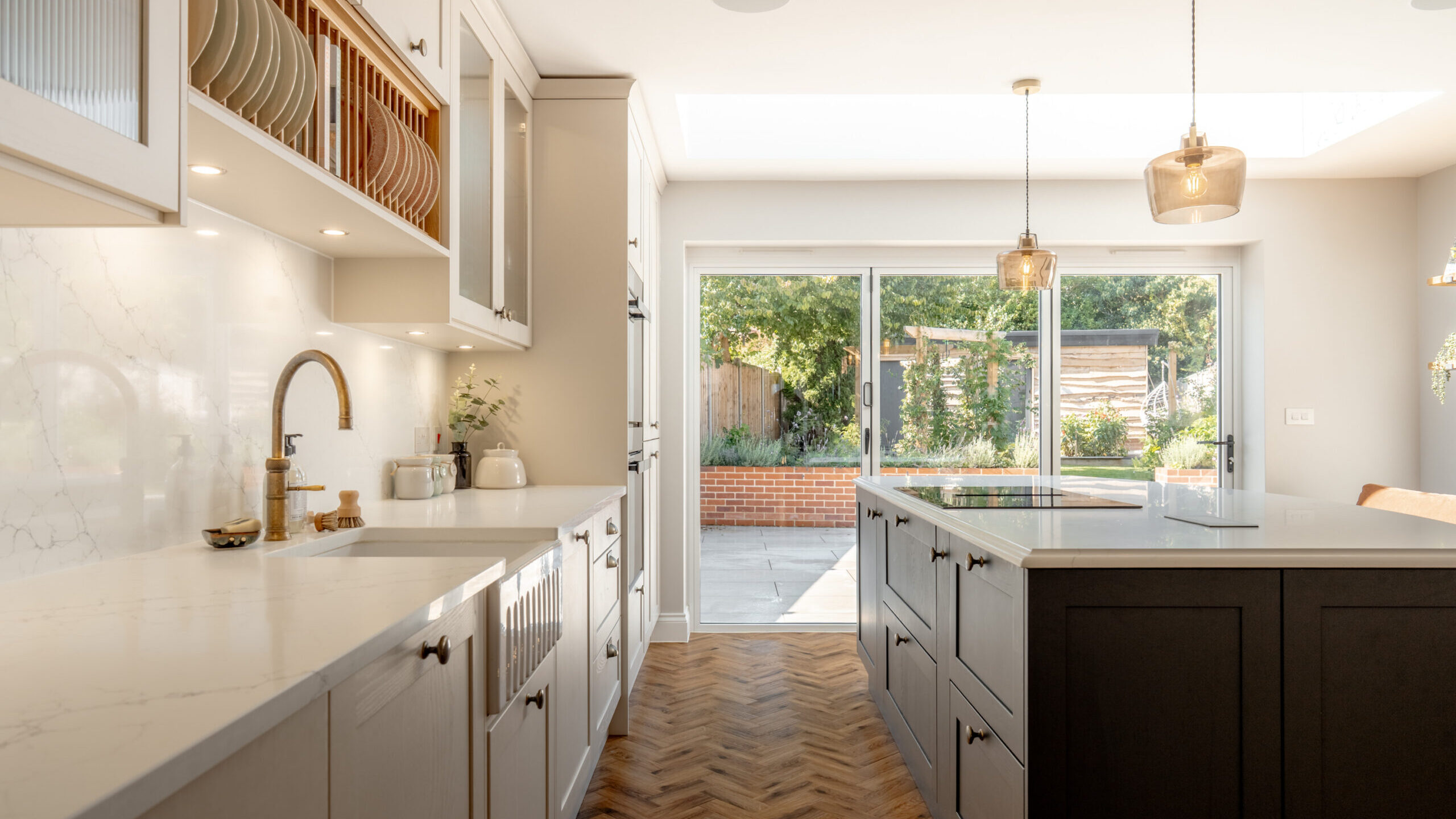 Modern kitchen with white cabinets, marble countertops, a farmhouse sink, and an island with dark drawers. Large glass doors open to a sunny patio and garden with greenery outside. Pendant lights hang over the island.