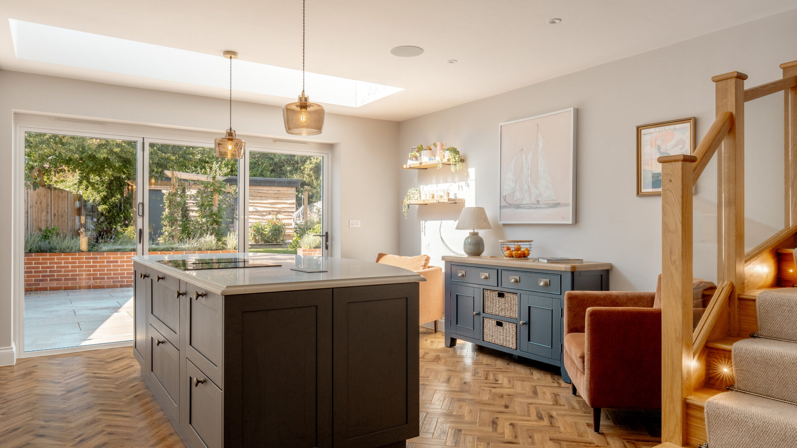 Bright modern kitchen with a dark island, wooden herringbone floor, and large sliding glass doors opening to a patio. There are two armchairs, a blue sideboard, wall art, and wooden stairs in the corner.