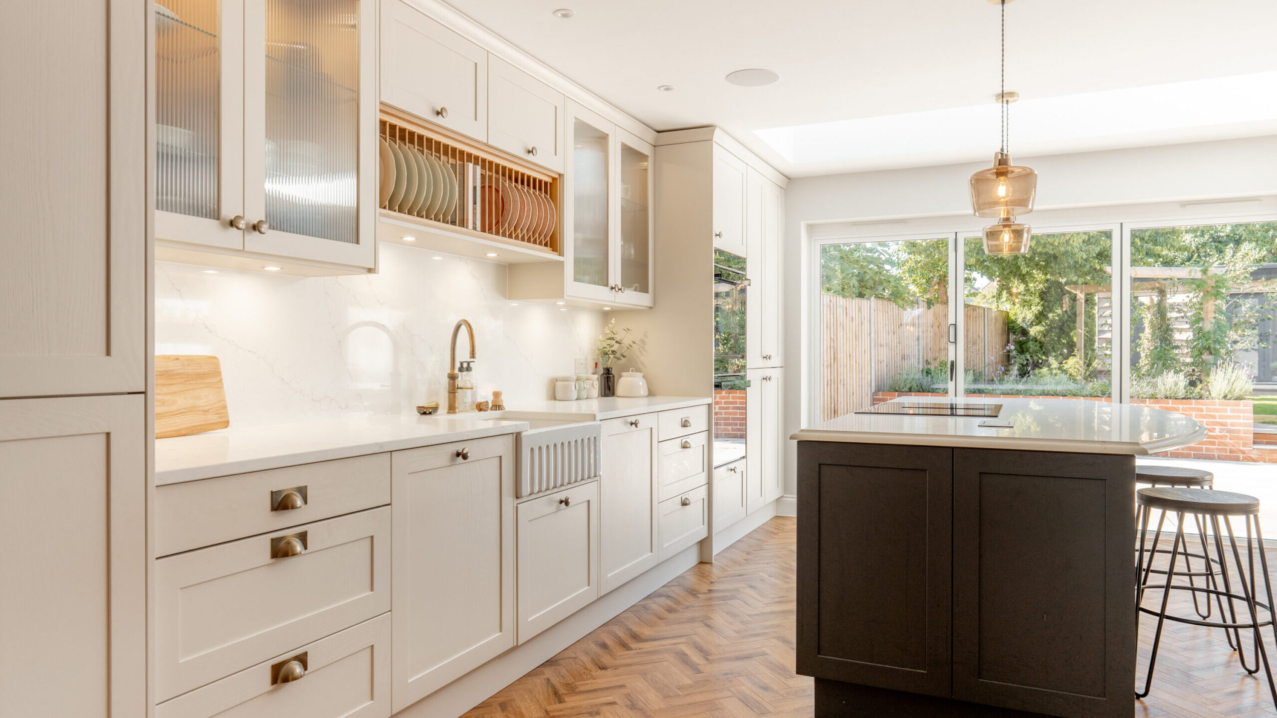 Modern kitchen with white cabinets, gold hardware, marble backsplash, and wood herringbone floor. A black island with barstools sits under a pendant light. Large glass doors open to a green backyard.