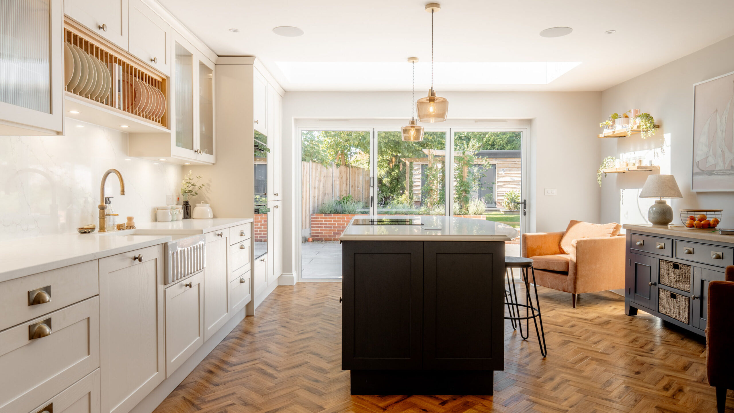 Bright, modern kitchen with white cabinets, a dark island, brass fixtures, and wooden herringbone floor. Large glass doors open to a garden. There’s a seating area with an armchair and sideboard on the right.