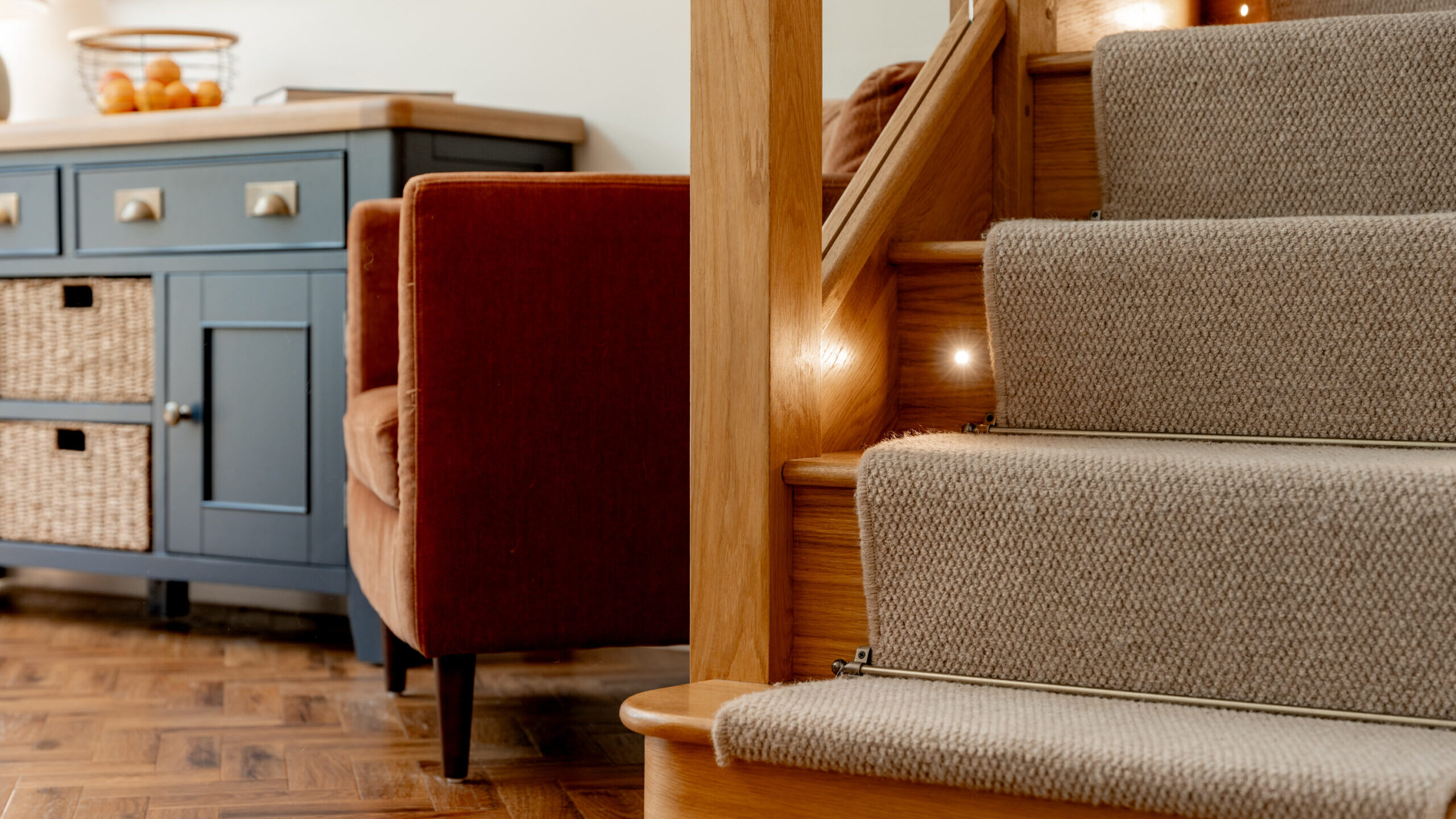 Close-up of carpeted wooden stairs with subtle stair lights, next to a blue sideboard and an orange-brown armchair on a herringbone wood floor in a cozy home interior.