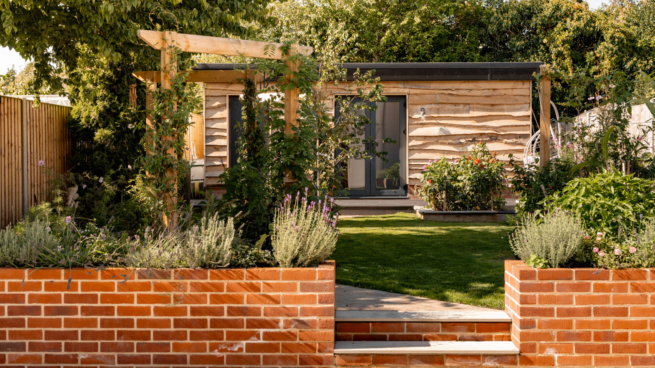 A small wooden garden house sits at the back of a lush garden, framed by trees and plants. A brick pathway with steps leads through raised brick flower beds toward the house.