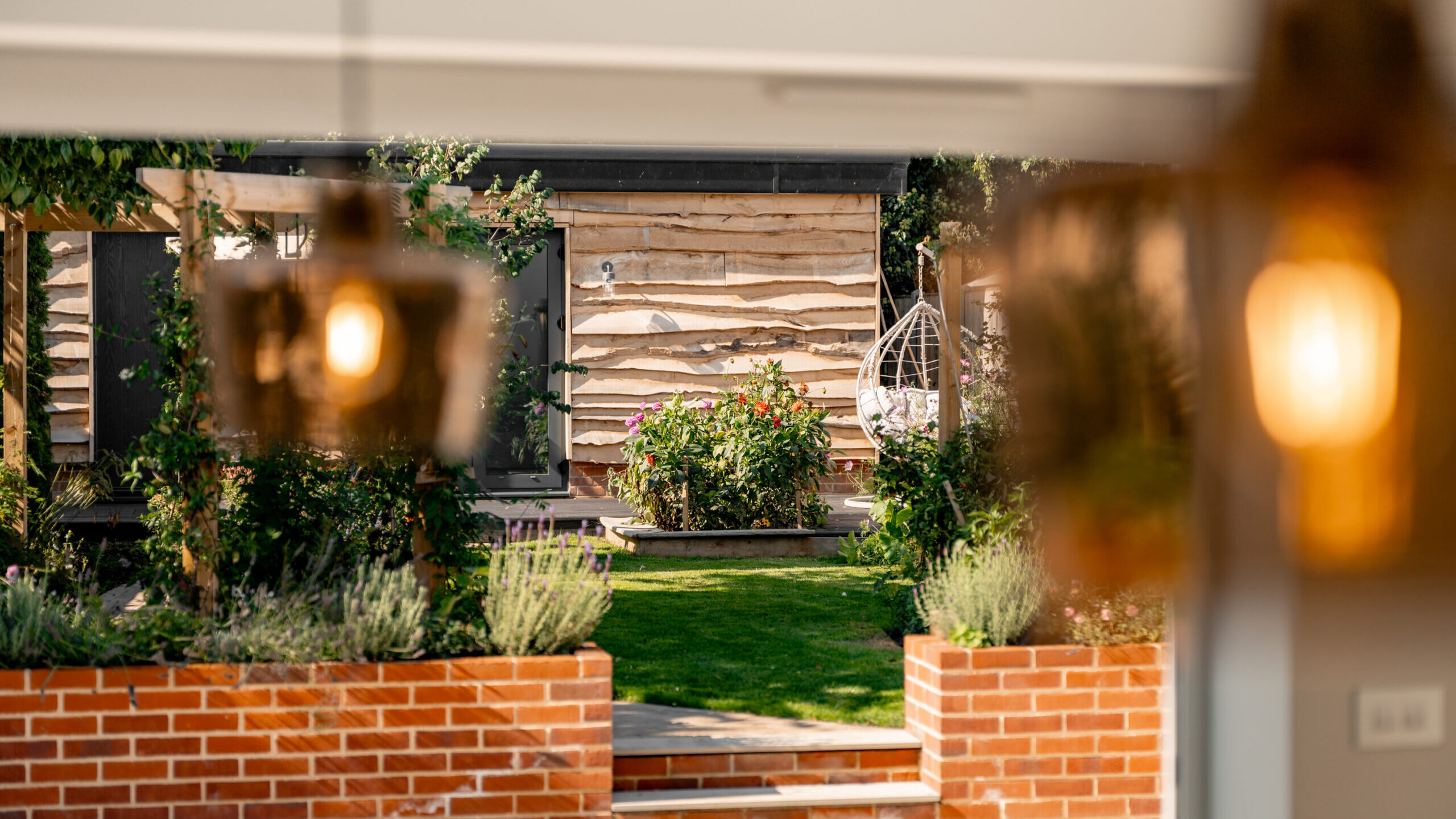 View of a garden with a manicured lawn, brick planters, and lush greenery, leading to a wooden building; blurred pendant lights are seen in the foreground.