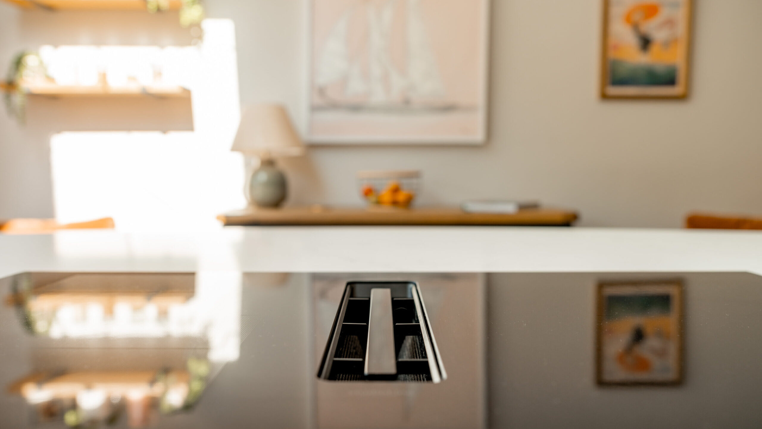 Close-up of a modern kitchen island with a built-in vent on a glossy surface. In the blurred background, there are shelves with plants, framed artwork, and a lamp on a sideboard.