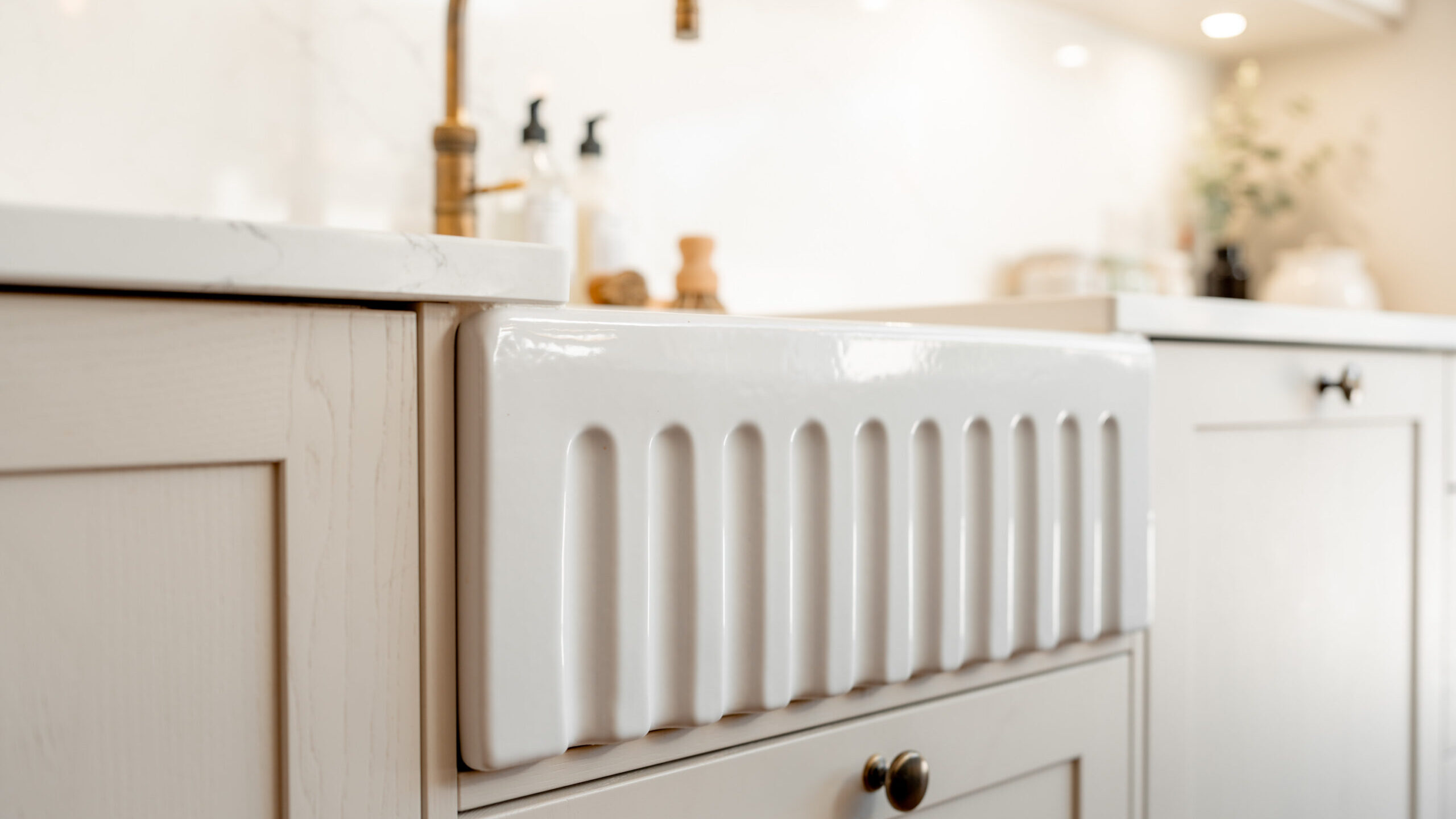 Close-up of a modern farmhouse-style white ceramic sink with vertical ridges, set in light-colored cabinetry and topped with a brushed brass faucet in a bright, minimal kitchen.
