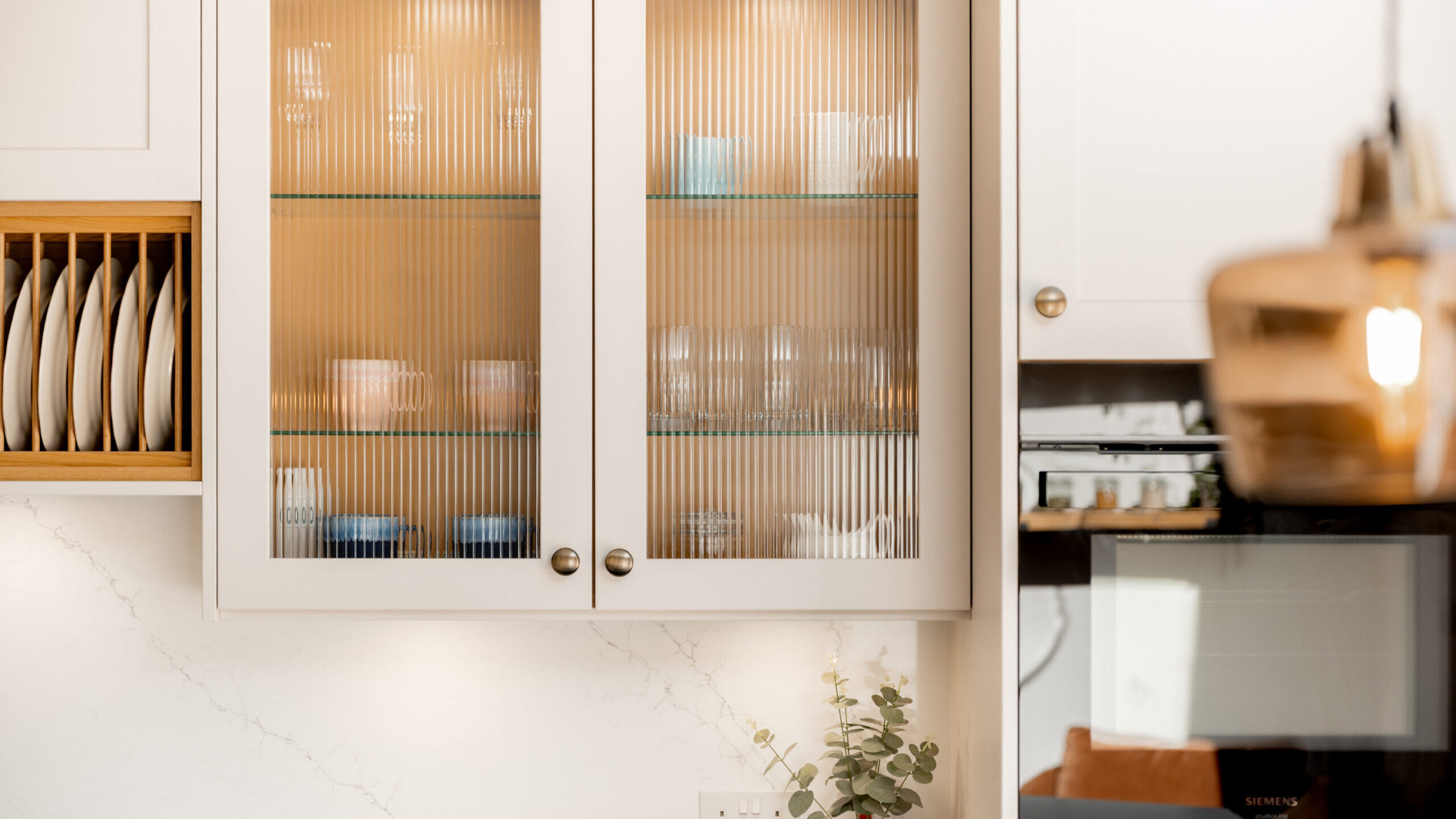 A modern kitchen with glass-front cabinets displaying neatly arranged cups and bowls, a plate rack with white dishes, a countertop with mugs and a small plant, and a built-in oven. Warm, soft lighting brightens the space.
