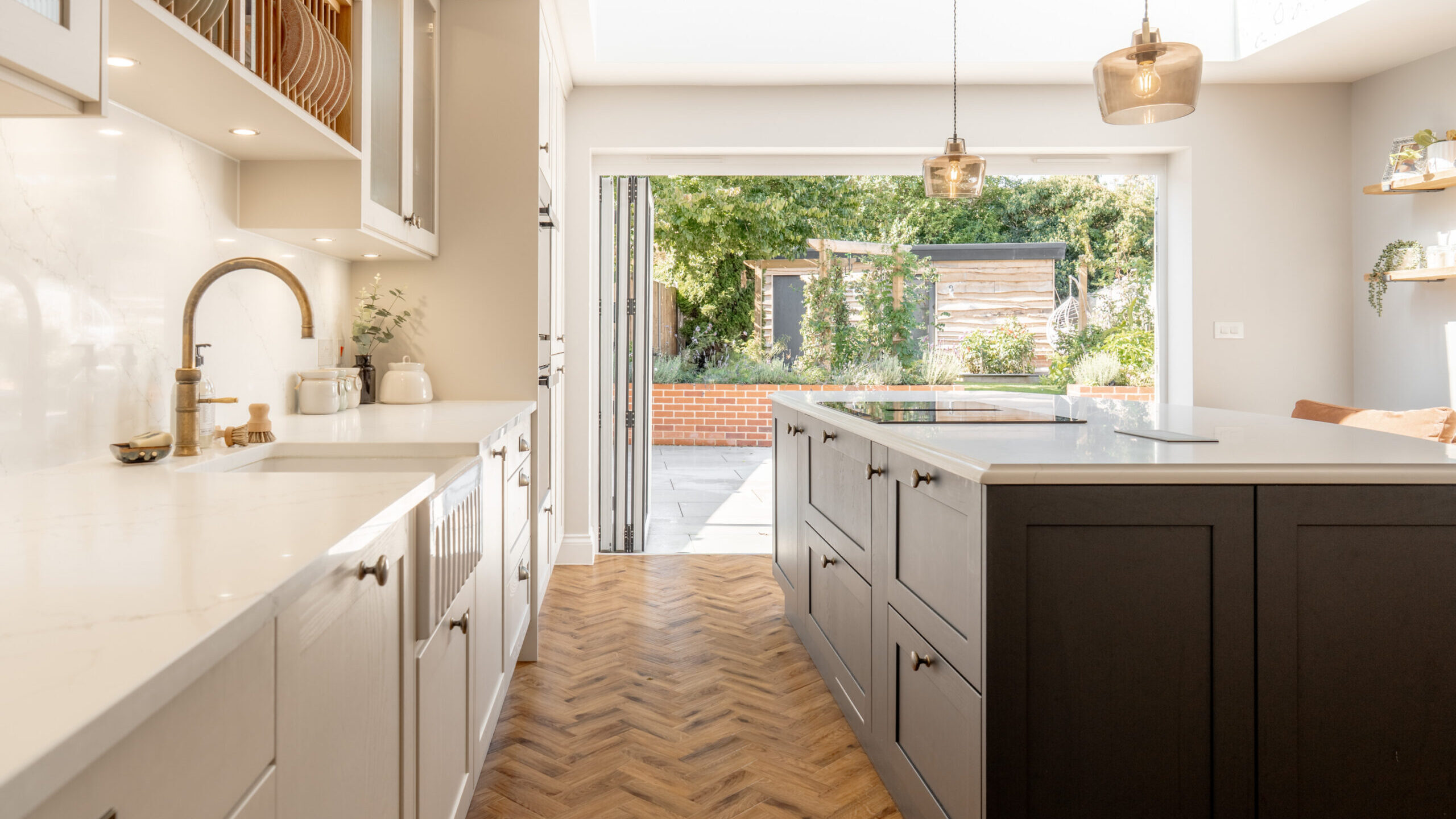 Modern kitchen with white cabinets, a dark island, and marble countertops. The space opens to a sunny patio and garden through large glass doors, and features wood herringbone flooring and pendant lighting.