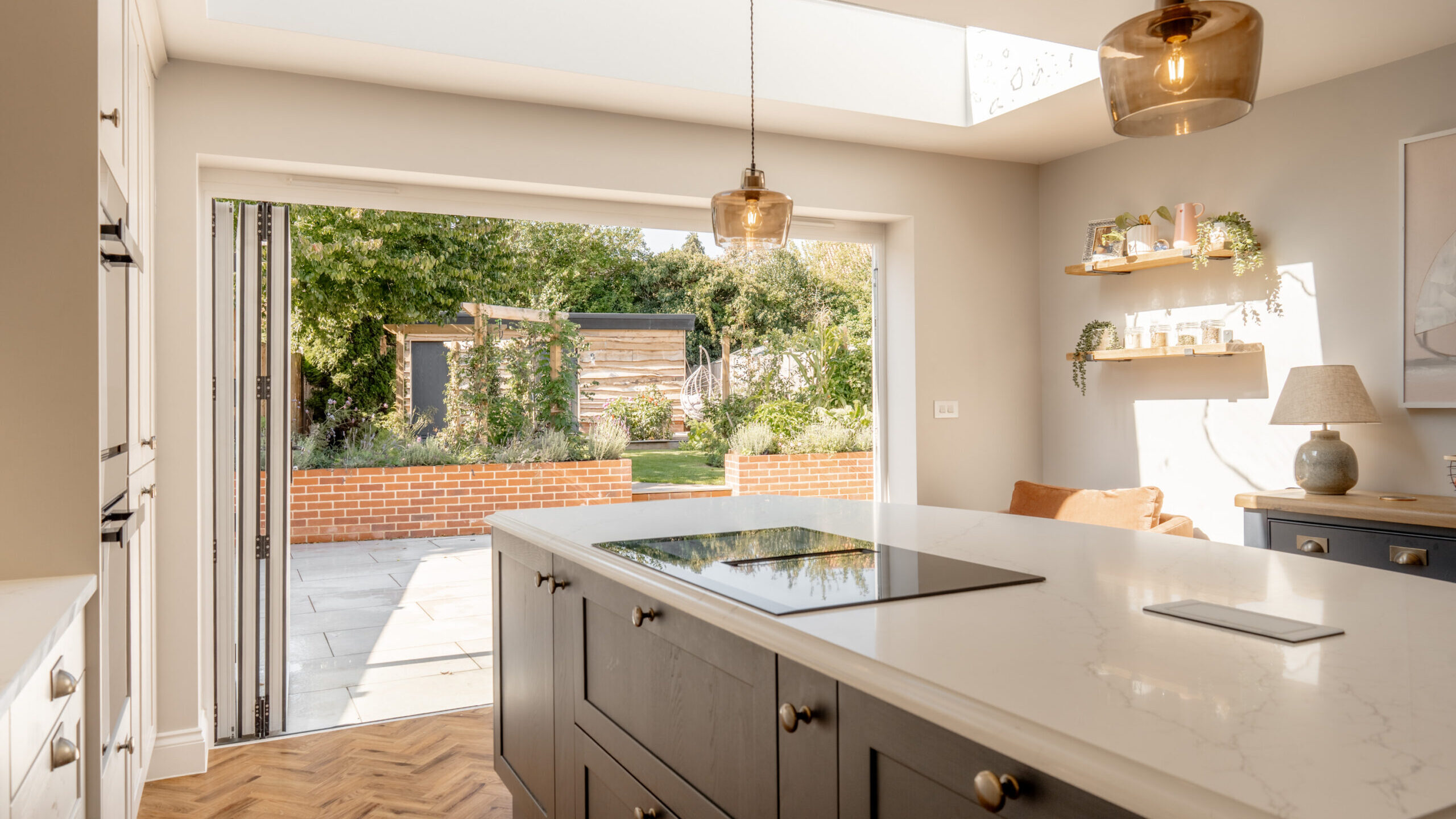 Modern kitchen with a marble island and induction cooktop, large folding glass doors open to a sunny garden with a shed, brick wall, and patio; pendant lights and open shelving enhance the bright, airy space.