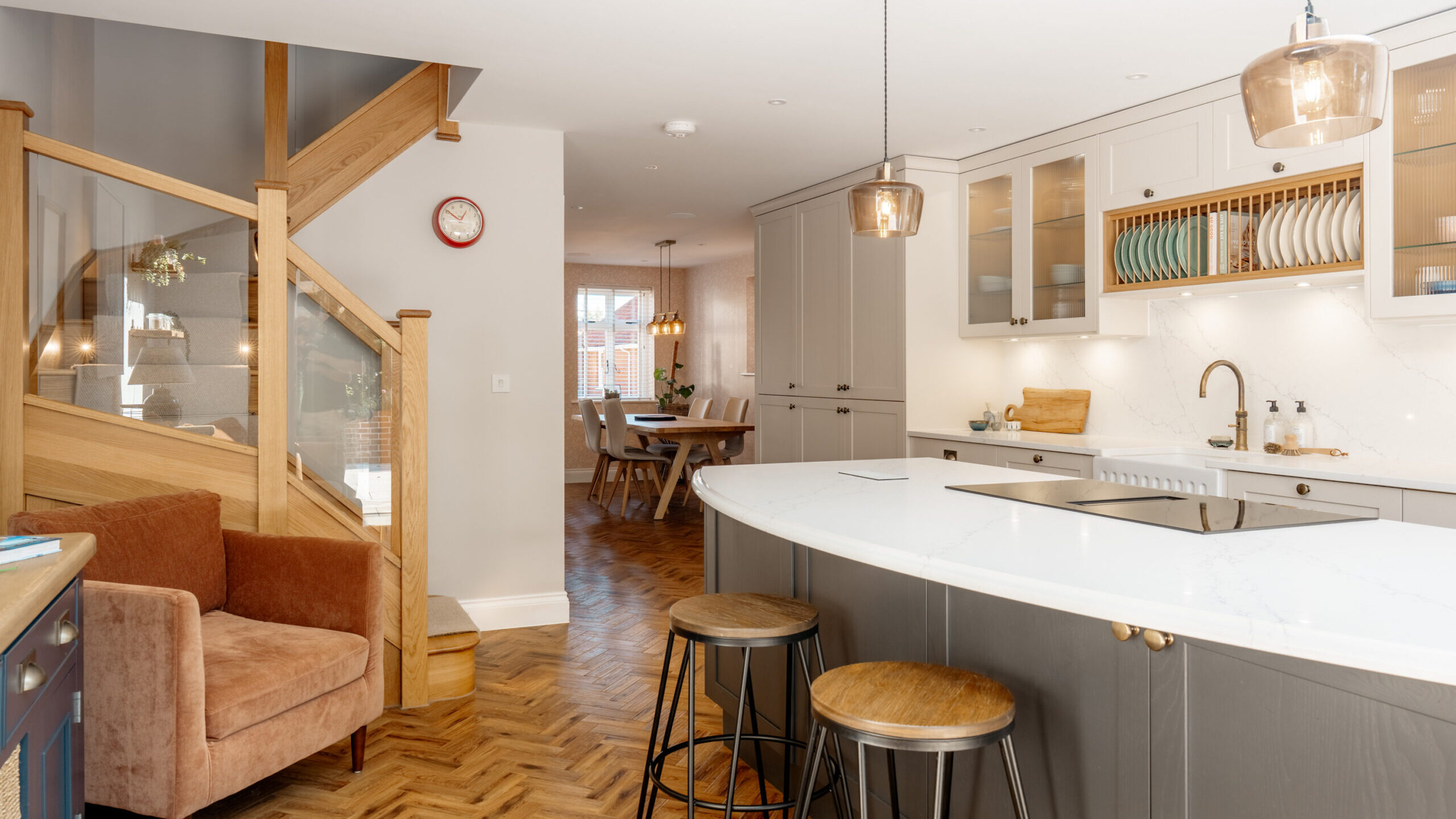 Modern kitchen with gray cabinets, a curved white island with two wooden stools, pendant lights, and a brown armchair near a wooden staircase; dining area visible in the background.