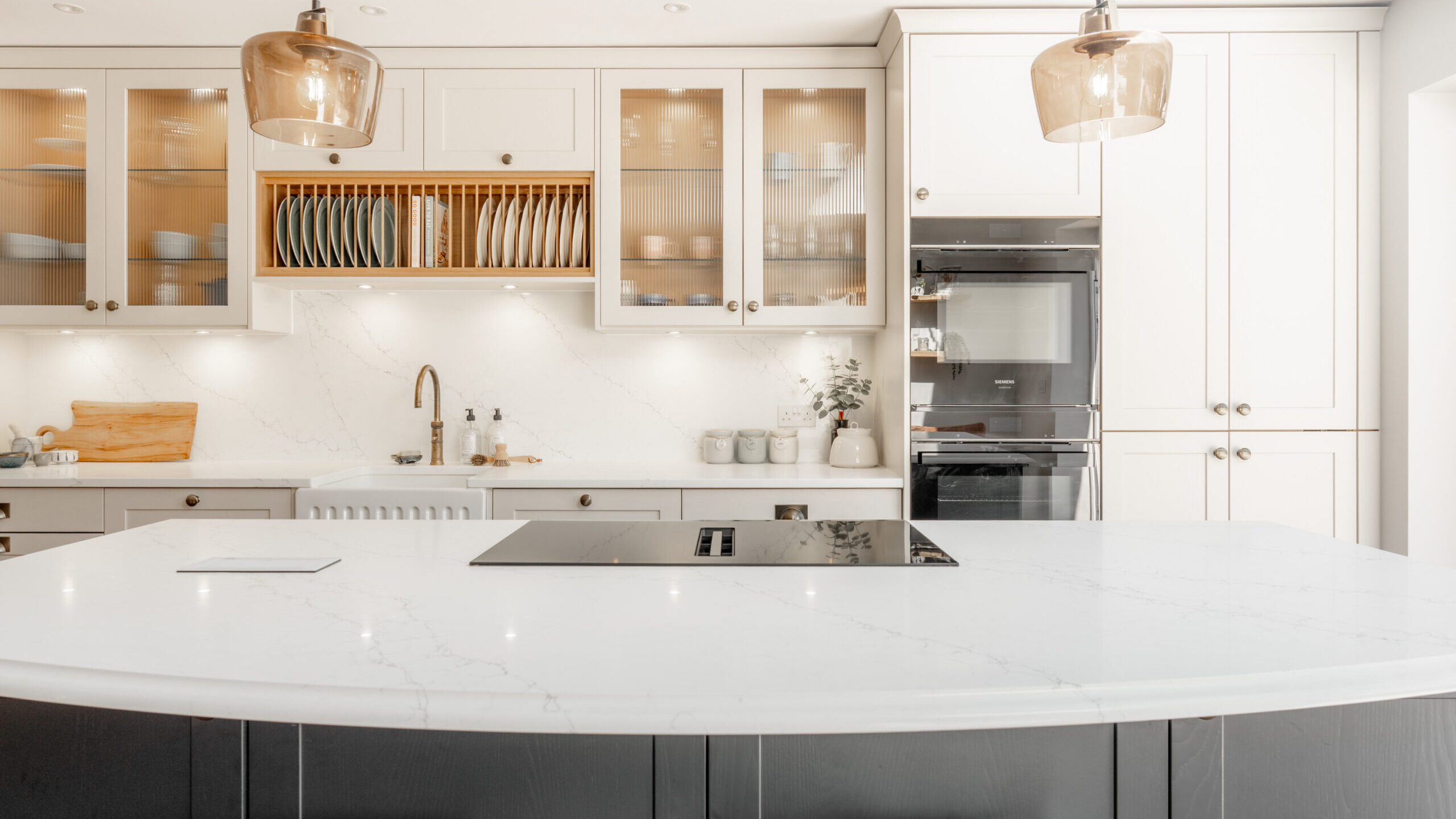 Modern kitchen with a white marble island, built-in stovetop, wooden stools, pendant lights, glass-front cabinets, open shelving for dishes, and integrated appliances against light cabinetry.