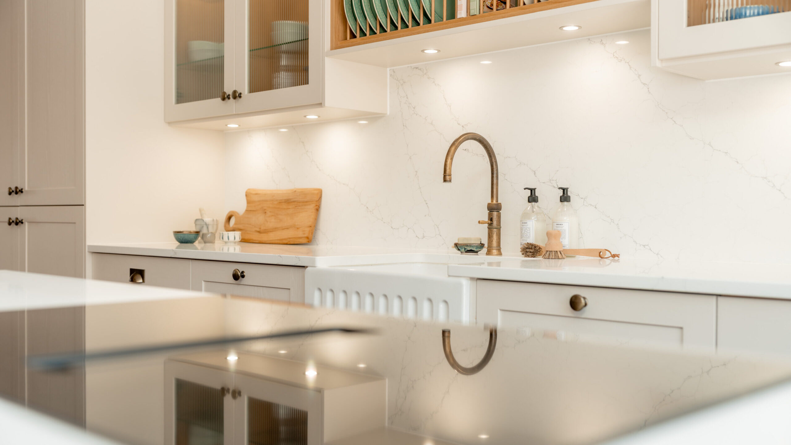 Modern kitchen with white cabinets, marble backsplash, a bronze faucet, dish soap bottles, and a wooden cutting board on the counter. Glass-front cabinets display dishes and bowls above the sink area.
