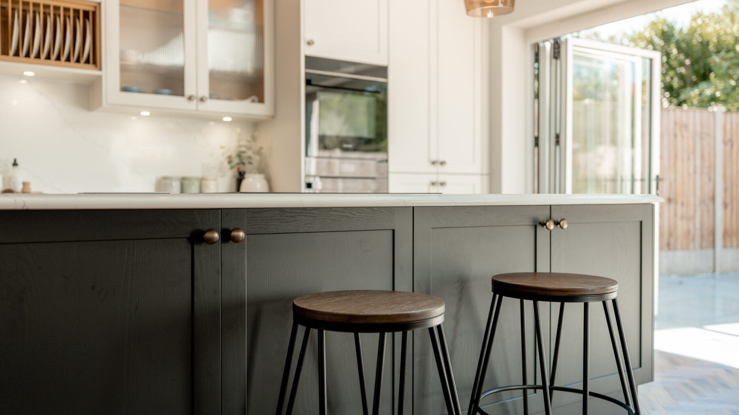 Modern kitchen with a dark island featuring two wooden bar stools. Light cabinets, glass doors, and natural light from open glass doors create a bright, inviting space with a view of the backyard.