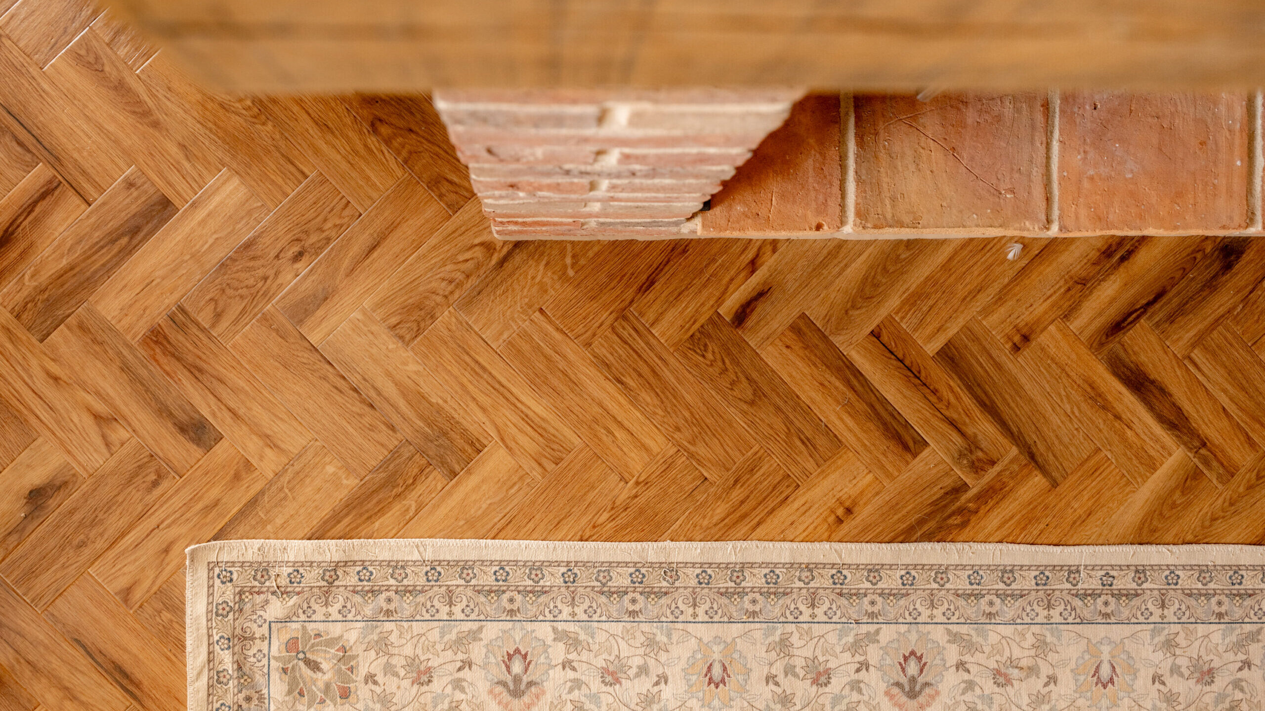 A top-down view of wooden herringbone-pattern flooring, a brick and tile structure, and the corner of a cream-colored patterned rug.