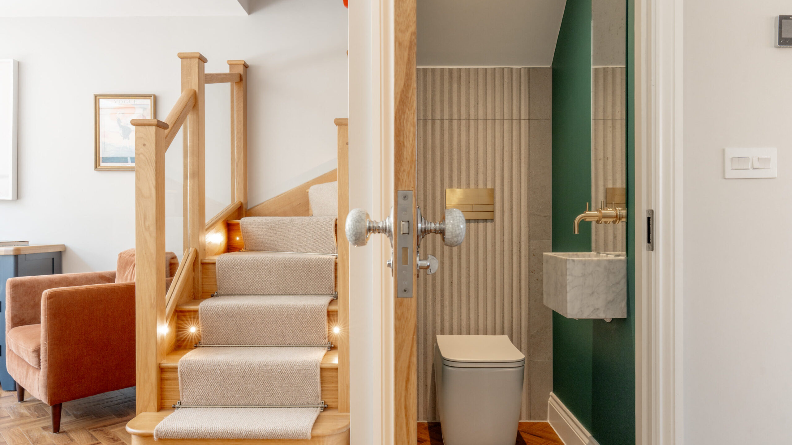 A view of a small bathroom with a modern toilet and sink next to a staircase with beige carpet and wooden handrails. A brown armchair is visible in the adjacent room.
