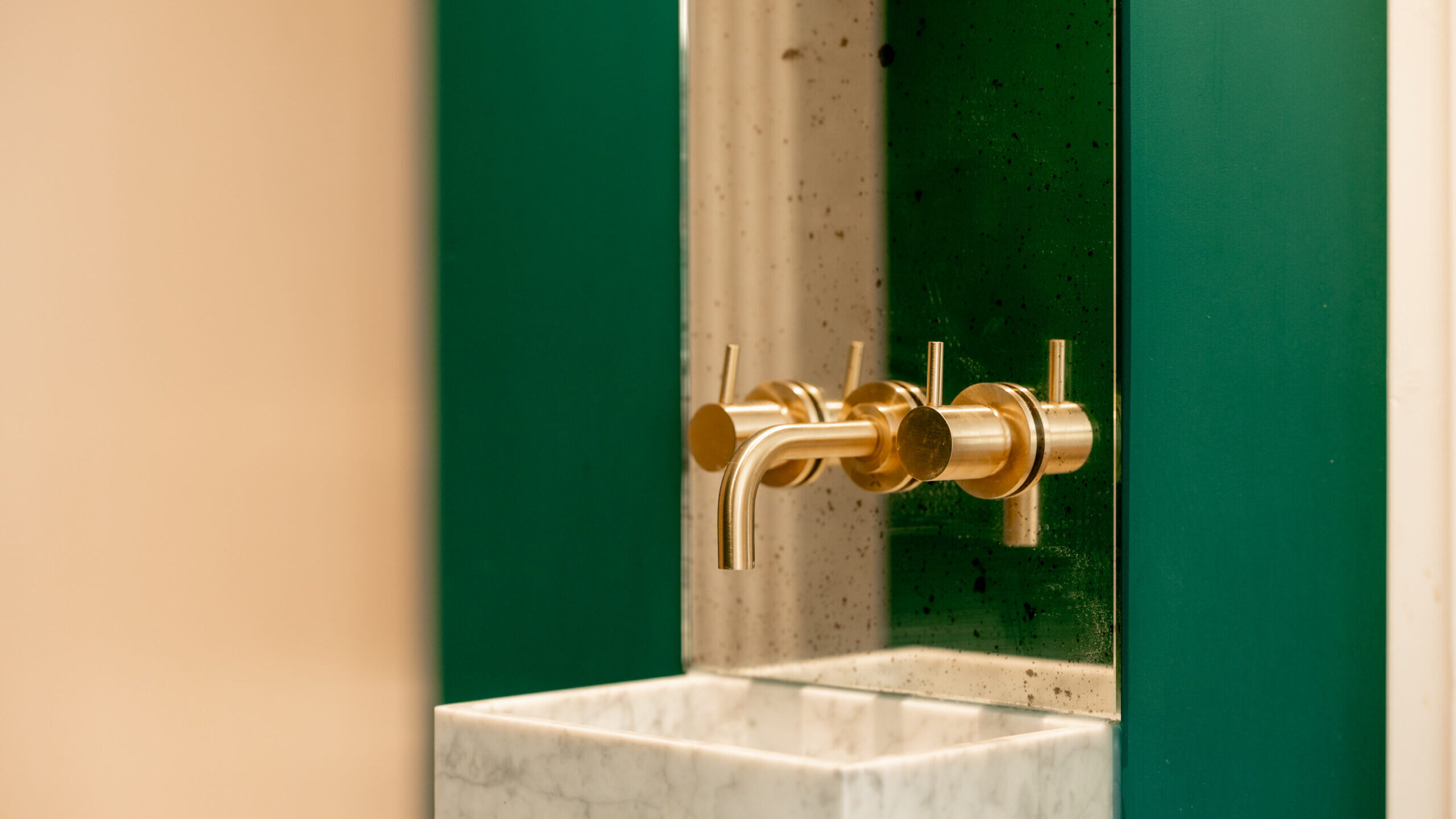 Close-up of a modern bathroom sink with a rectangular white marble basin, gold wall-mounted faucet and handles, and a green glass backsplash. The design is minimalist and contemporary.