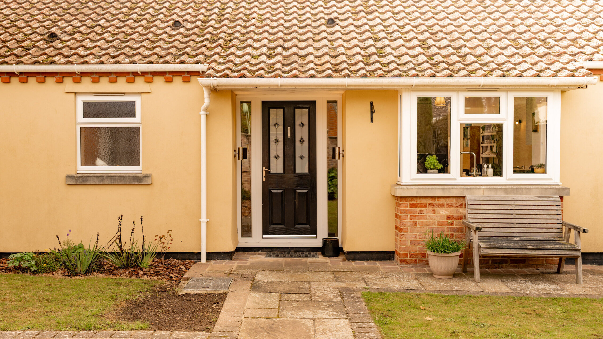 The image shows the front of a single-story house with cream-colored walls, a black front door, a white framed window, a wooden bench, a potted plant, and a paved pathway leading to the entrance.