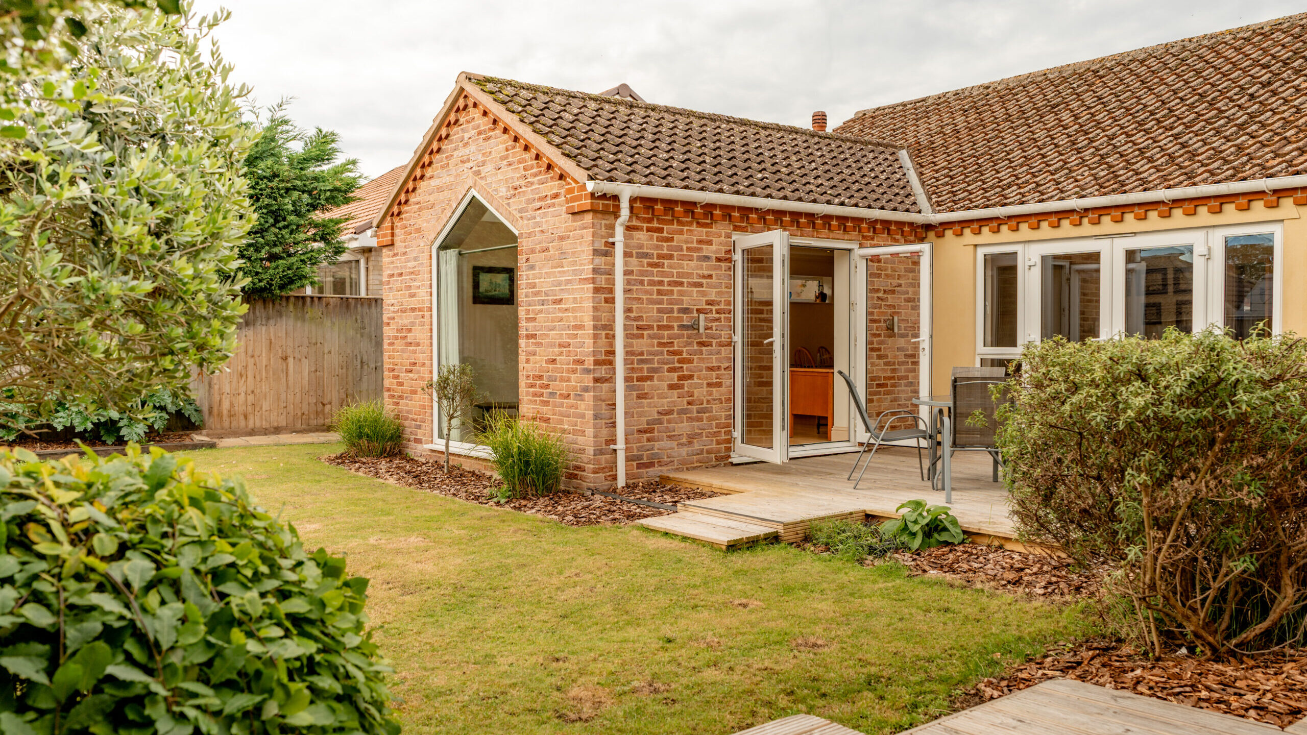 A red-brick house with a tiled roof, glass patio doors open to a wooden deck with outdoor chairs and table, surrounded by a well-maintained garden with grass, shrubs, and trees.